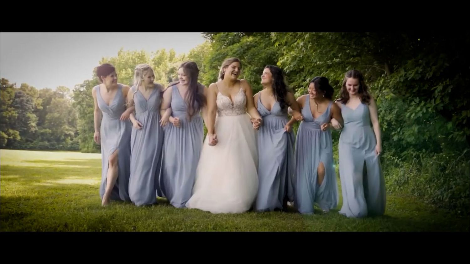 Bride in white gown laughing with bridesmaids in blue dresses outdoors on a sunny day.