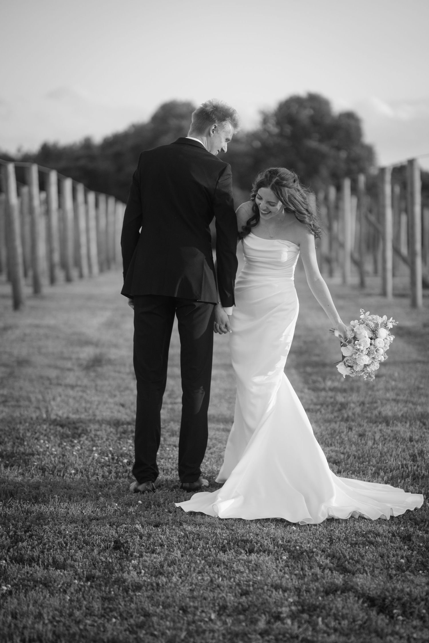 Black and white photo of bride and groom walking through vineyard, bride holding bouquet.