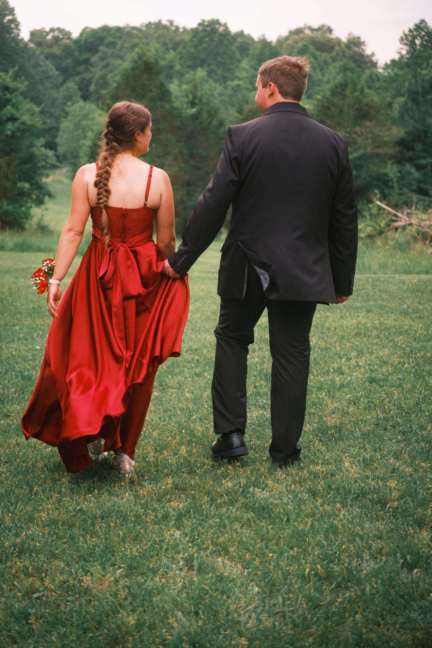 Couple walking hand-in-hand through green field, woman in red satin gown with braid, man in black suit, viewed from behind.