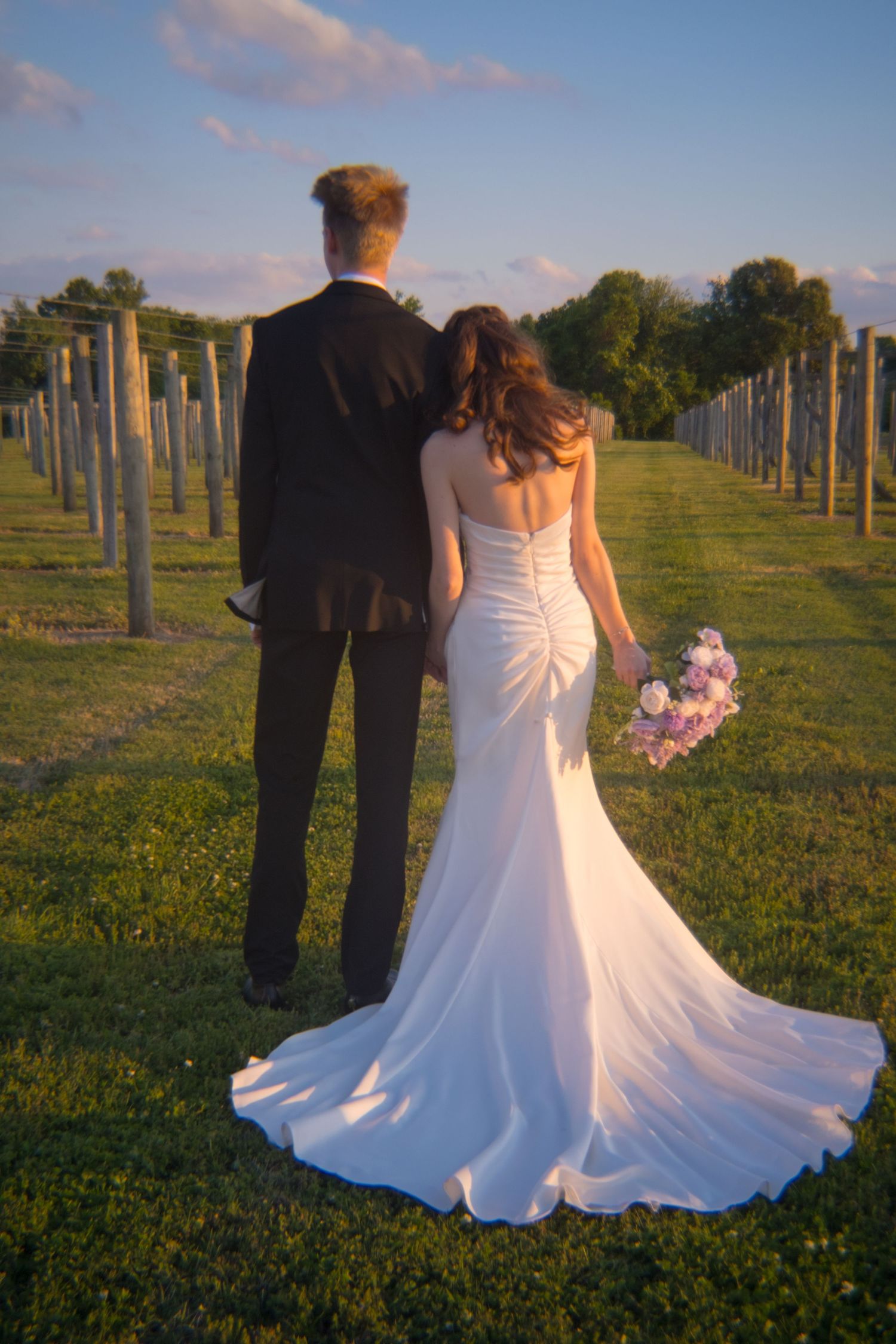 Bride and groom walking through vineyard rows at golden hour, bride in elegant white gown with train holding bouquet.