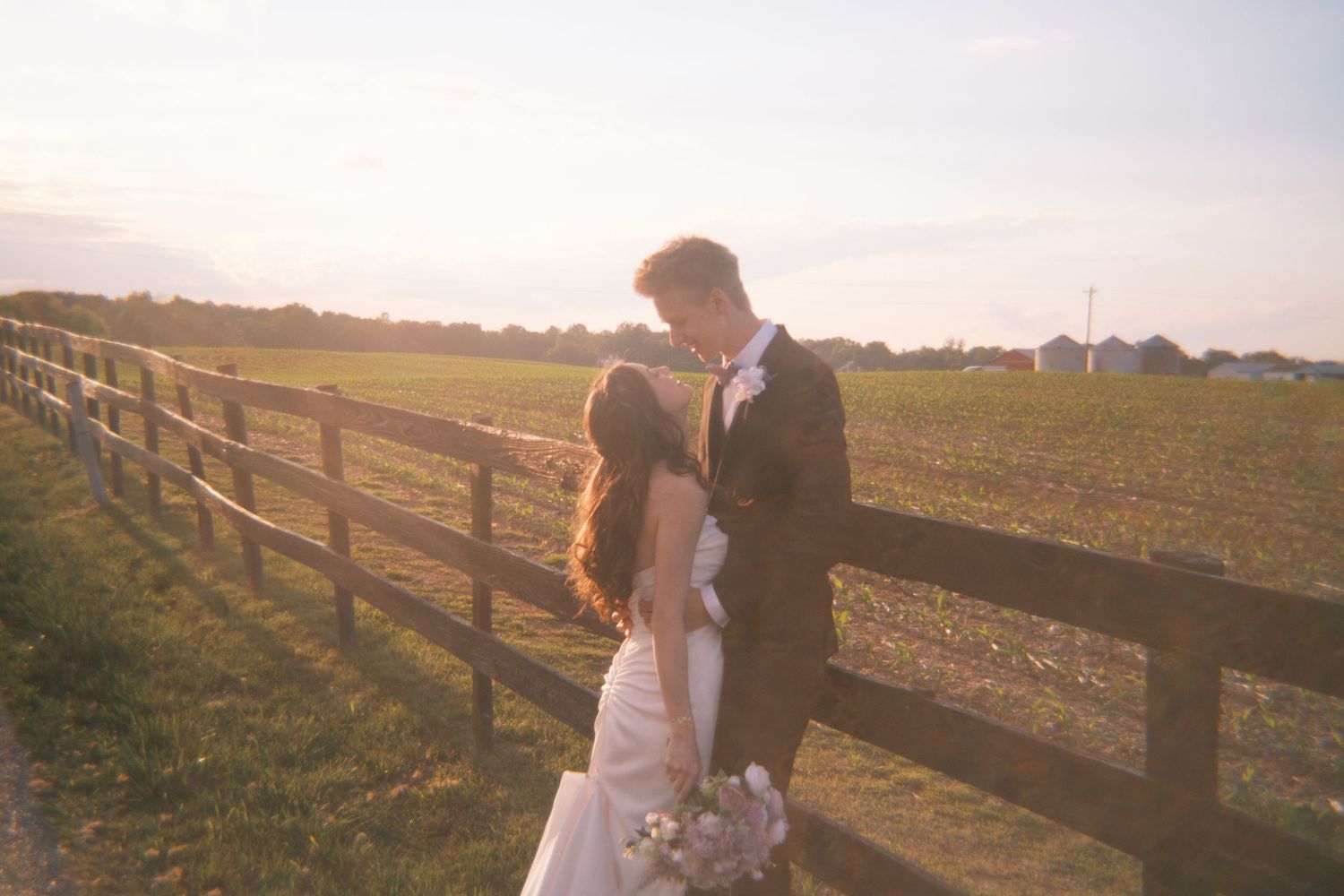 Bride and groom sharing a romantic moment by a wooden fence at golden hour on a farm.