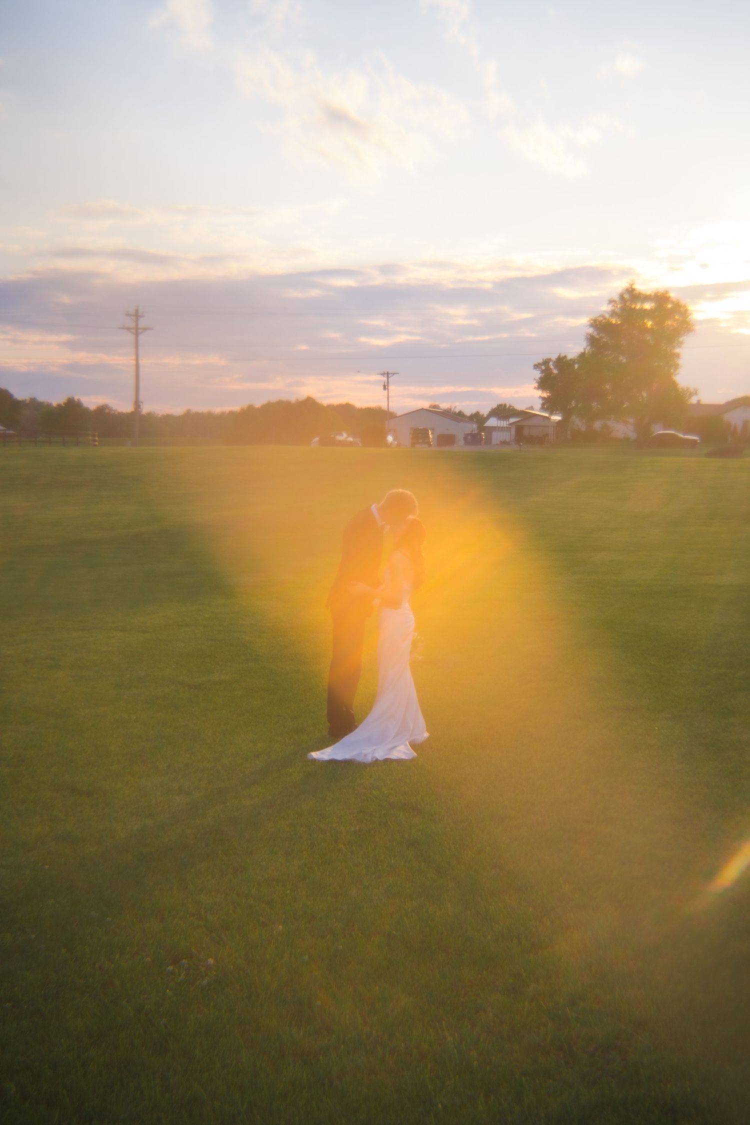 Bride and groom embracing at sunset on a green field with golden lens flare and pastel sky.