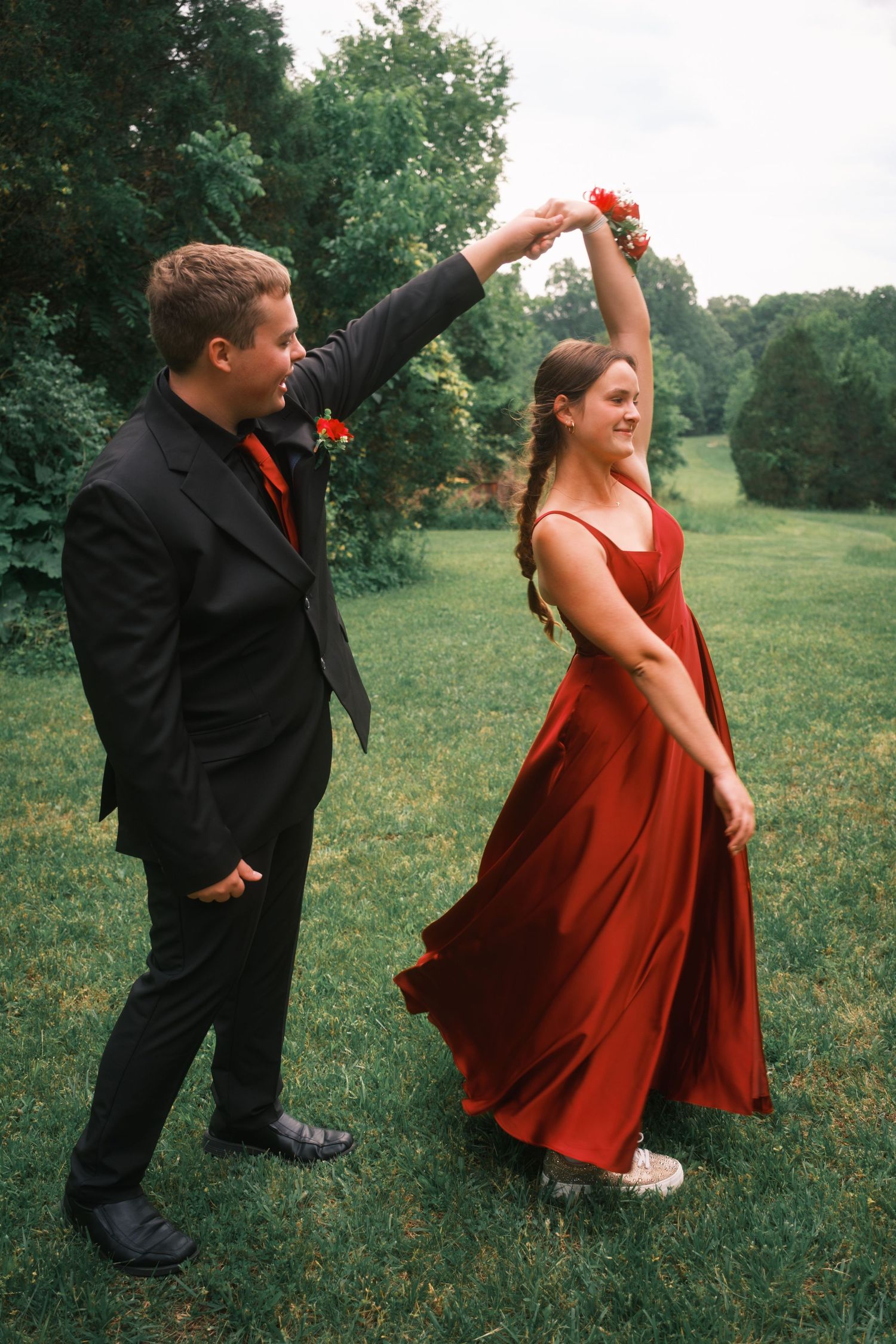 Young man in black suit spinning his date in a flowing red prom dress on green grass.