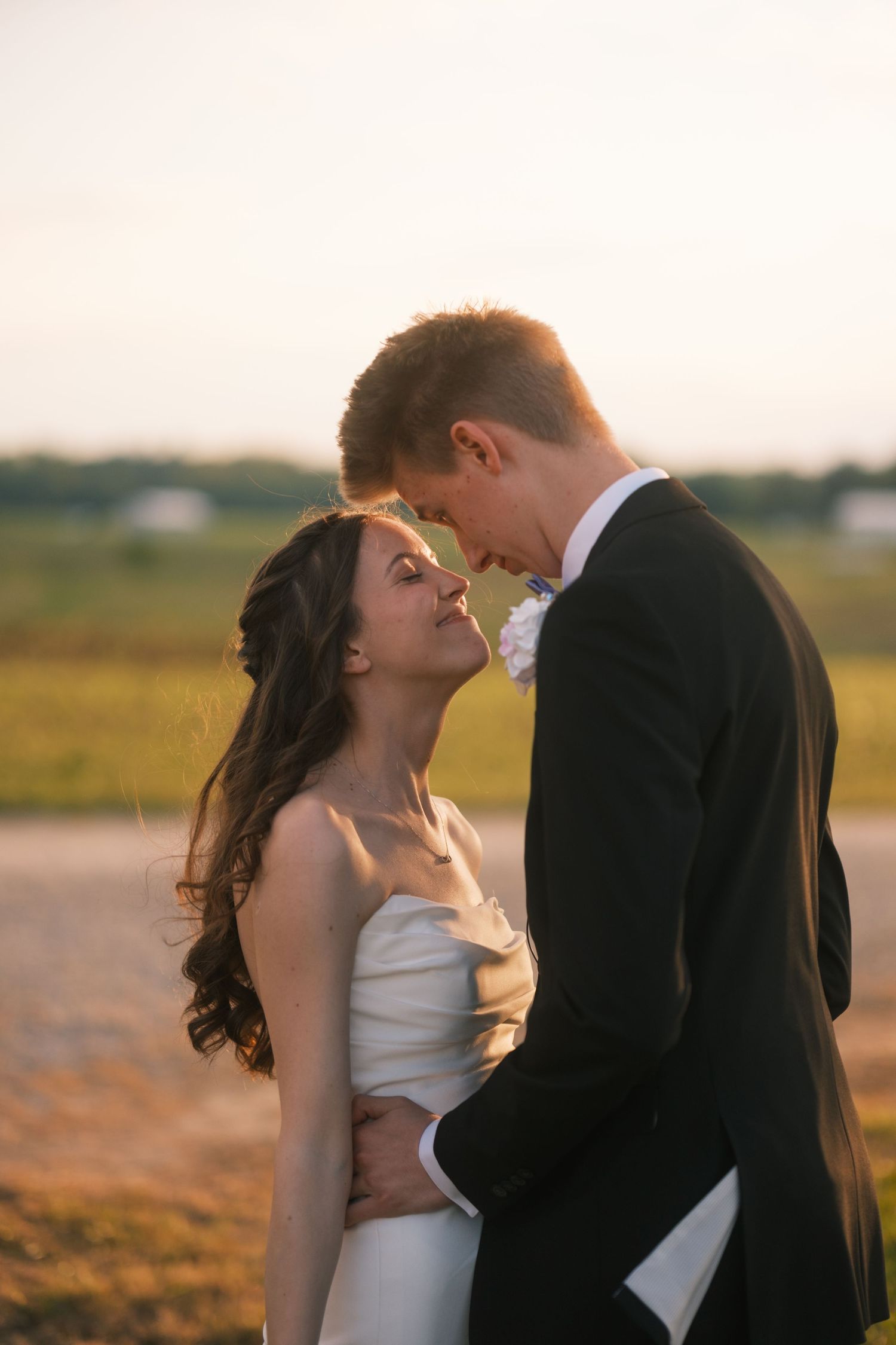A bride and groom share a romantic sunset moment, foreheads touching, as she wears a white gown and he a black tuxedo.
