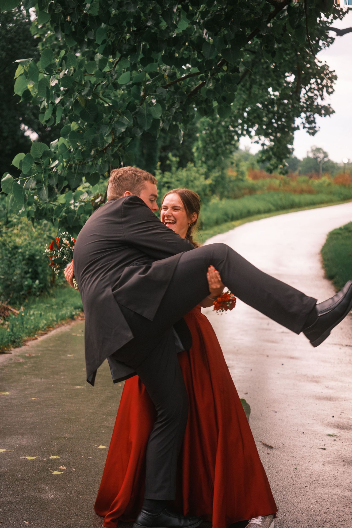 Groom lifting laughing bride in red dress outdoors on a rainy tree-lined pathway.