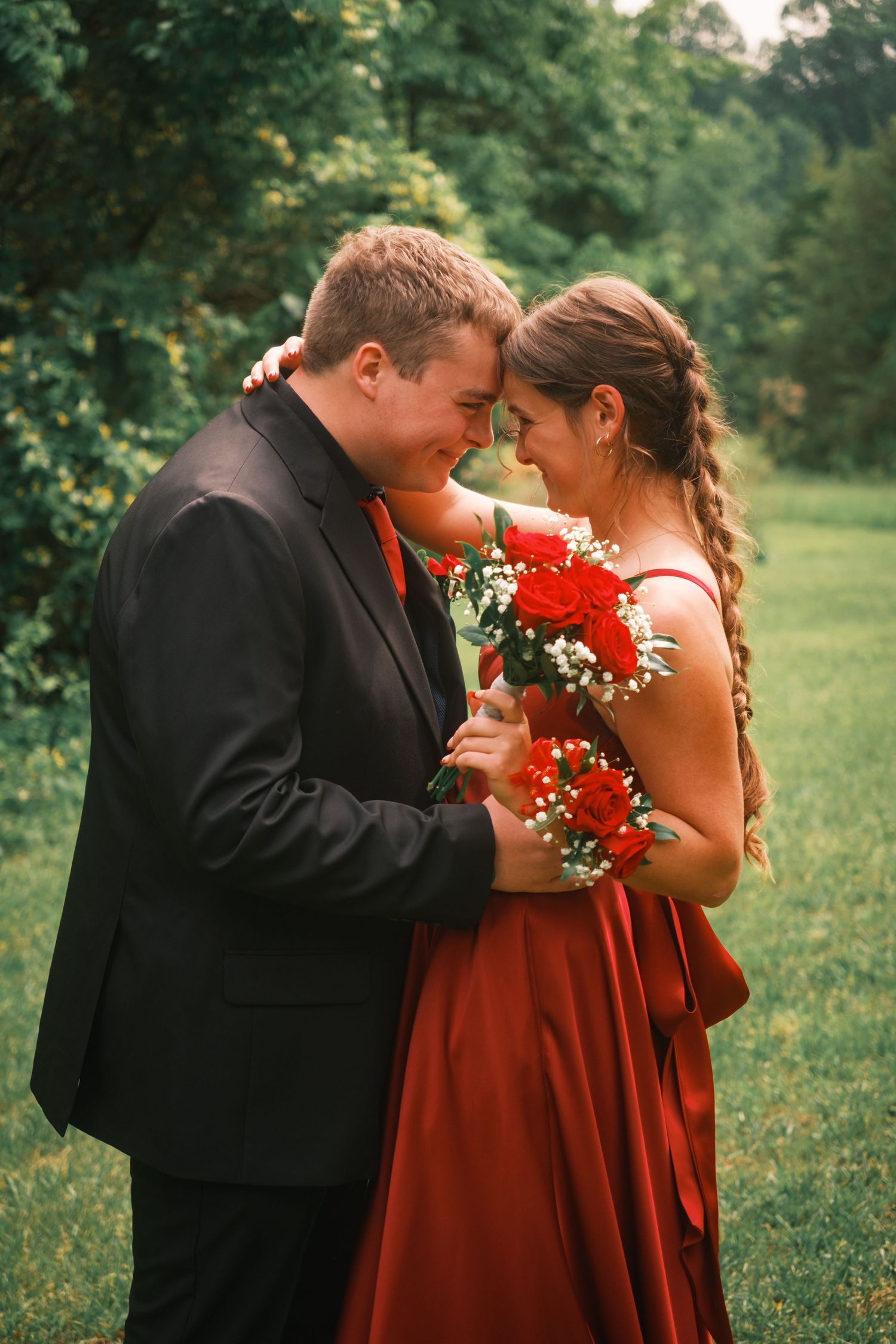 Prom couple in black suit and red gown touching foreheads while holding red roses outdoors.