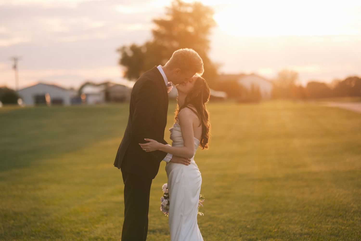 Bride and groom standing close together on a green lawn, sharing a tender moment at sunset.