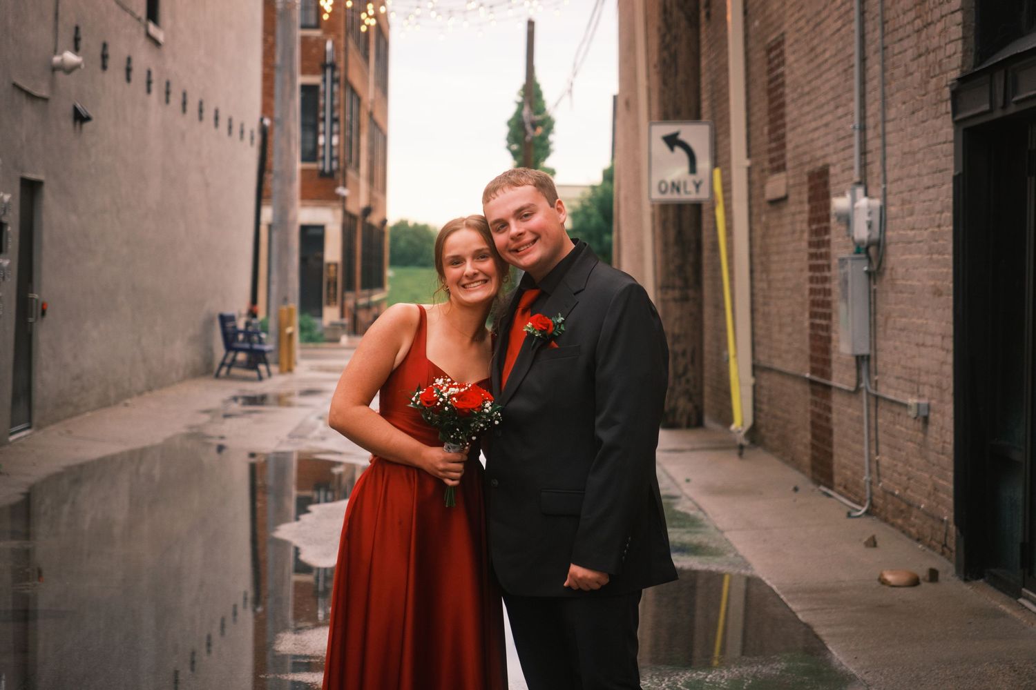Couple in formal attire posing in an alley, woman in red dress holding flowers, man in black suit with boutonniere.