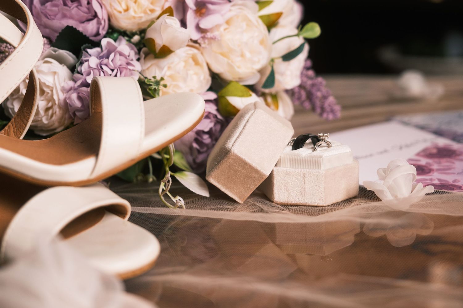 Elegant wedding flat lay with white heels, purple floral bouquet, ring box with diamond rings, and invitation card.