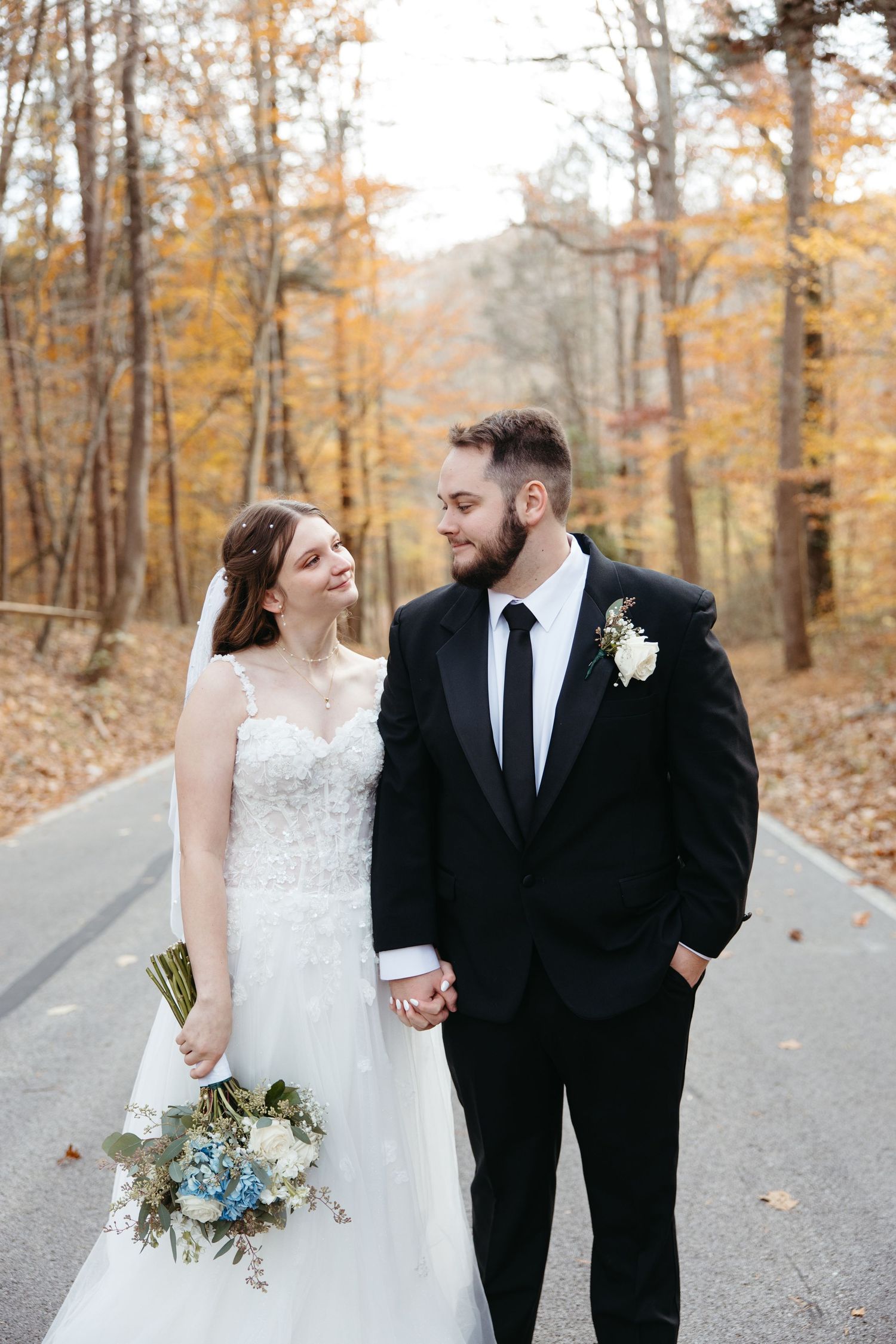 Bride and groom holding hands on a tree-lined road in autumn, bride holding blue and white bouquet.