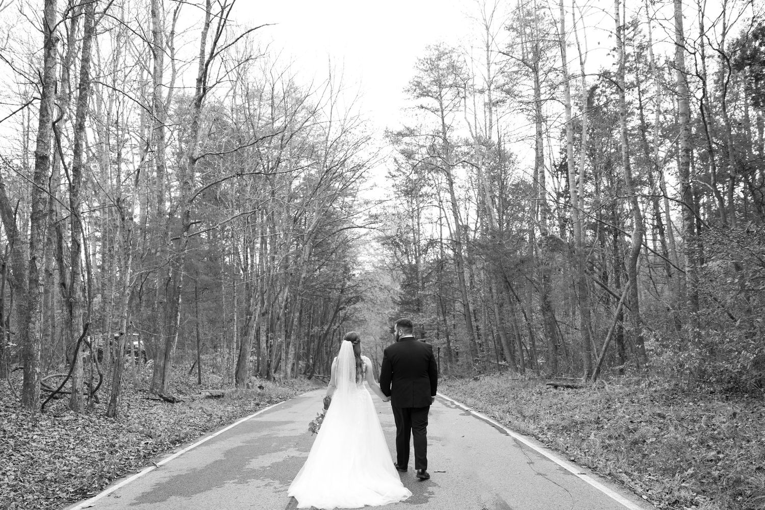Black and white photo of bride and groom walking hand in hand down a tree-lined path on their wedding day.