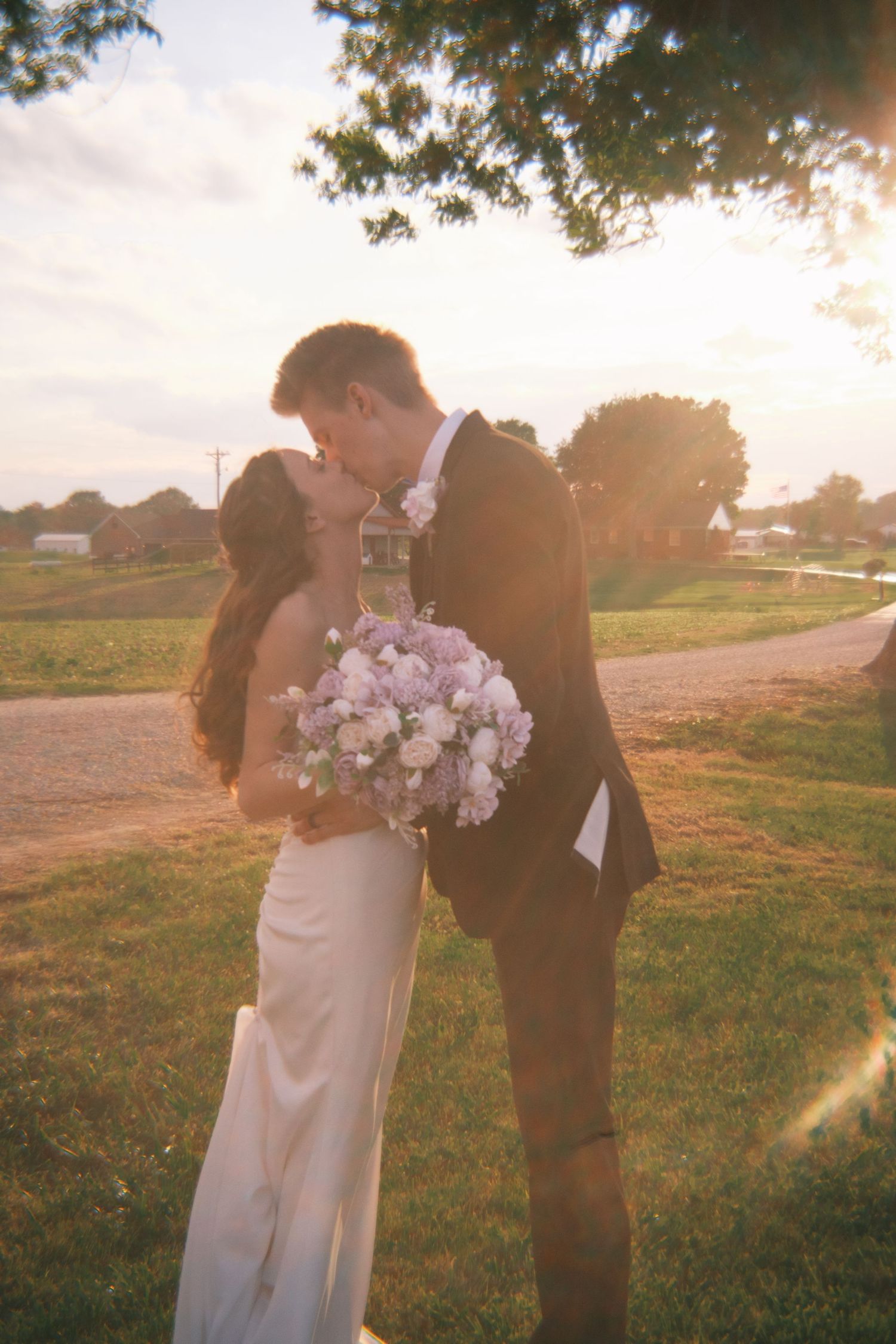Bride and groom sharing a kiss at golden hour, bride holding pink bouquet, sunlight glowing behind them.