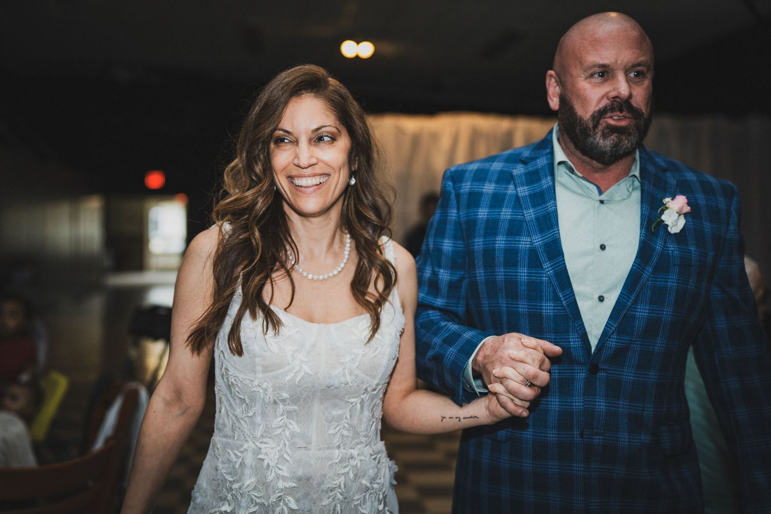 Bride in white lace gown and groom in blue plaid suit walk hand in hand at their wedding reception.