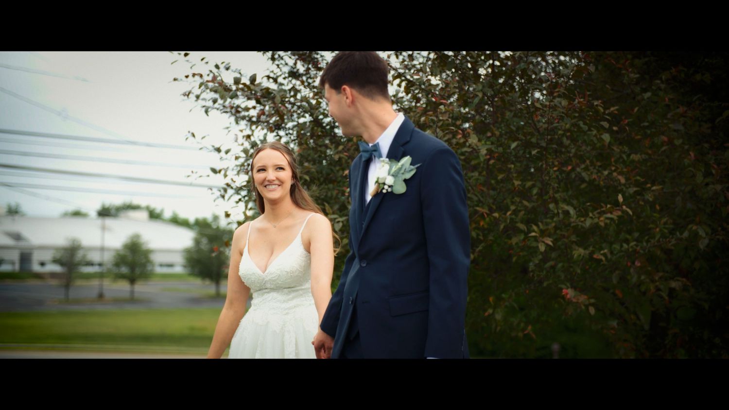 A bride in a white gown and groom in a navy suit hold hands outdoors near lush greenery on their wedding day.