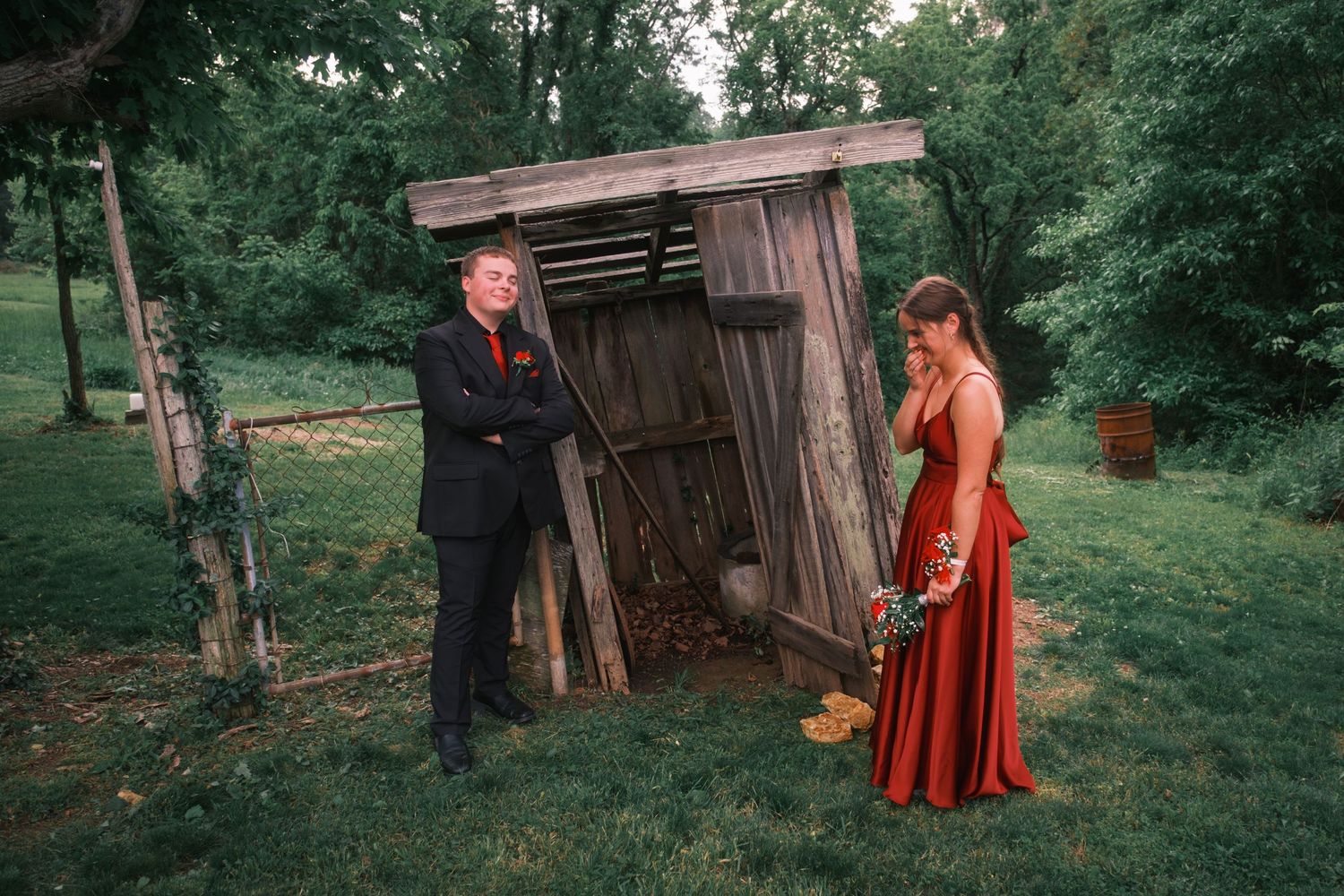 Prom couple posing beside a rustic wooden outhouse structure in a lush green outdoor setting.