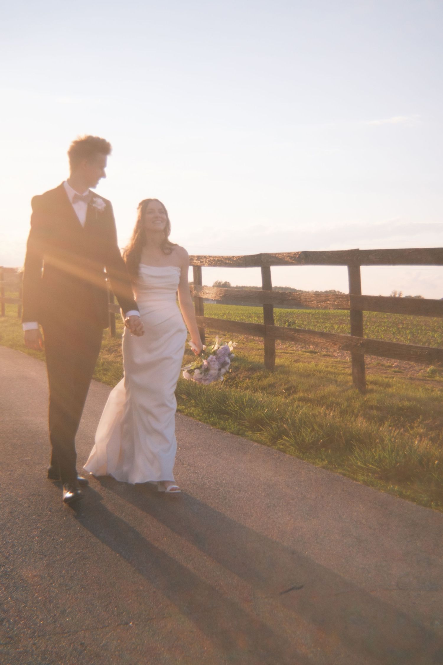 Bride and groom walking together along a fenced country road, bathed in warm golden sunset light.