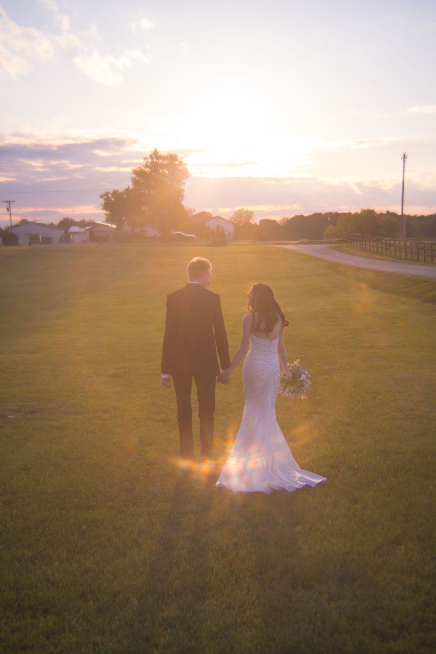 Newlyweds holding hands while gazing at a stunning sunset over a lush green field on their wedding day.