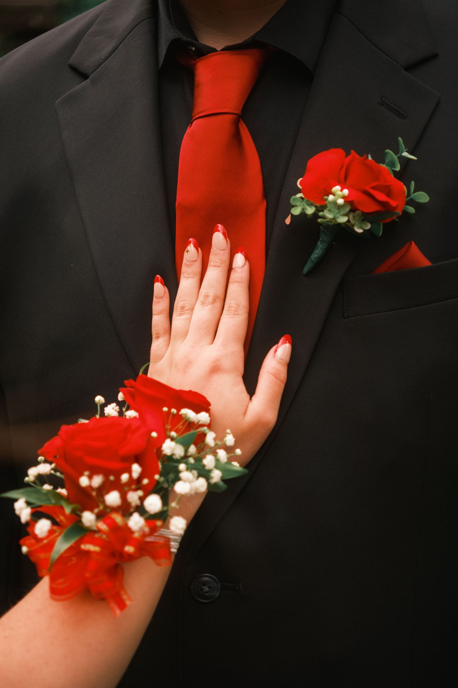Red rose wrist corsage on woman's hand resting on man's black tuxedo with matching red rose boutonniere and tie.