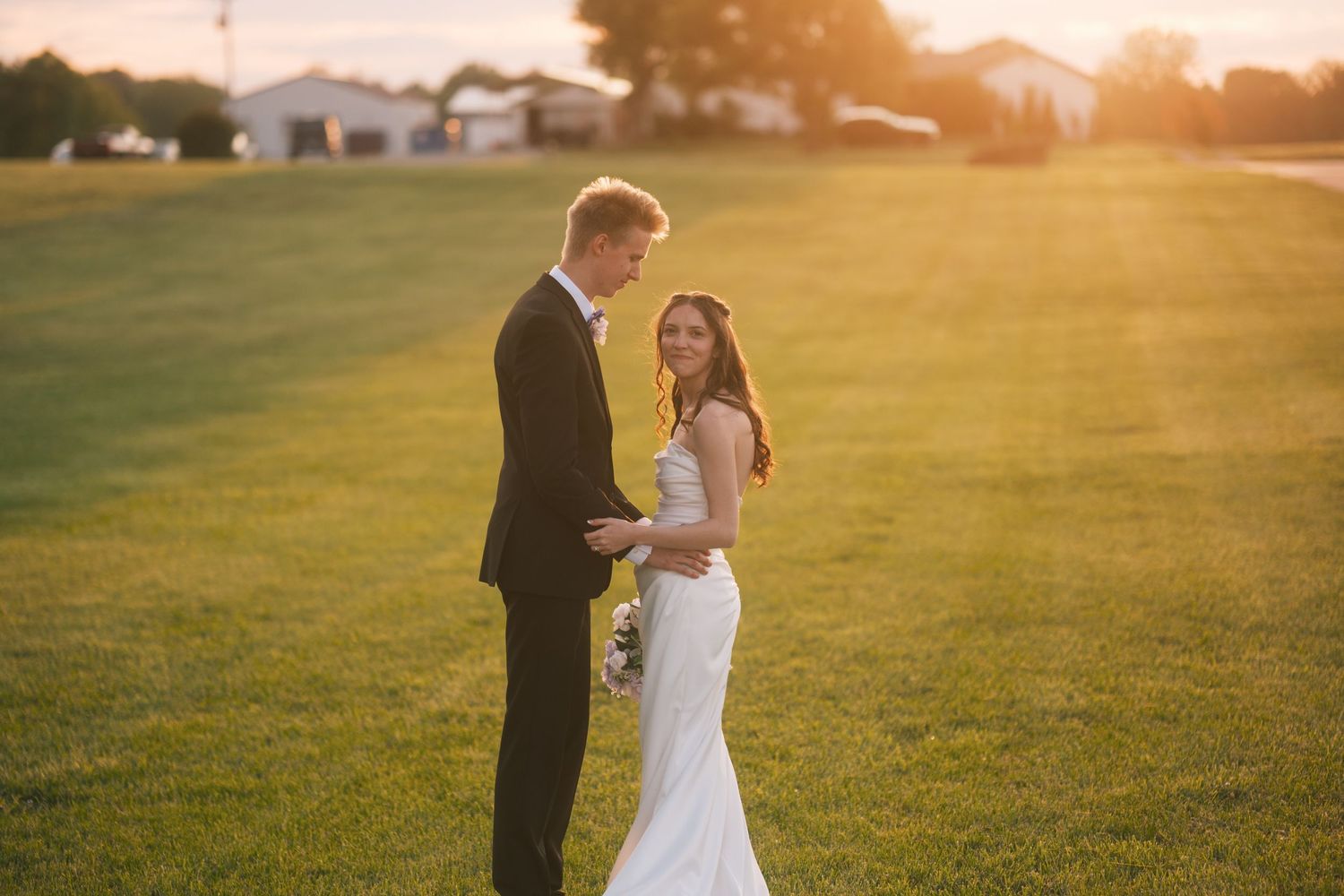 Bride in a fitted white gown and groom in a dark suit share a romantic moment at golden hour outdoors.