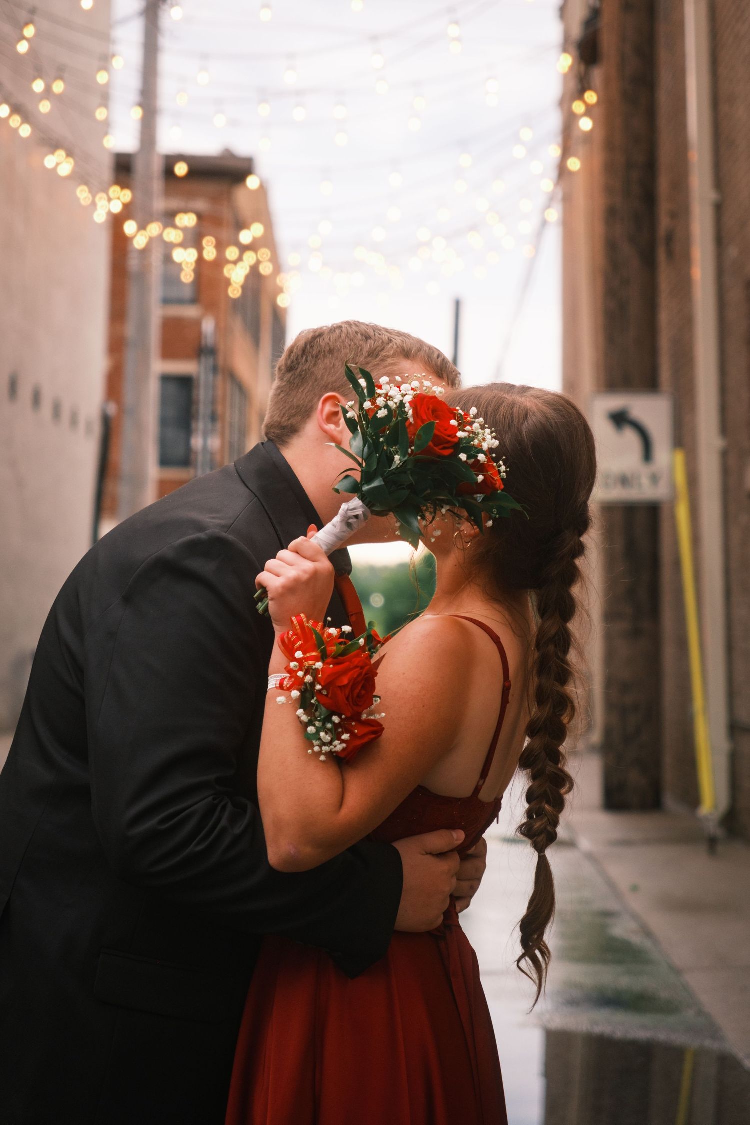 Couple kissing in an alley with string lights, woman in red dress holding floral bouquet with red flowers and baby's breath.