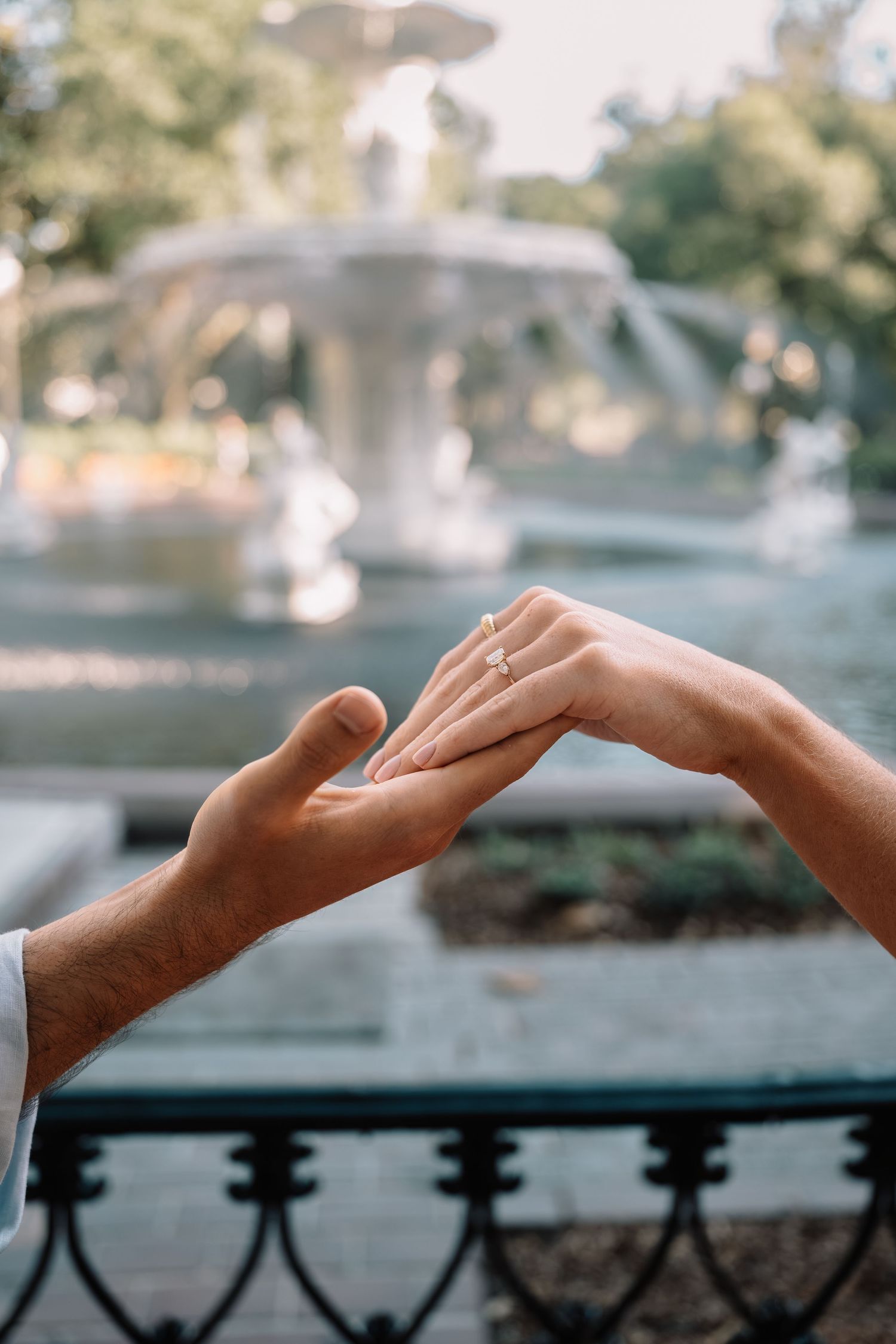 Close-up of engagement ring being placed on woman's finger near a fountain in a park.