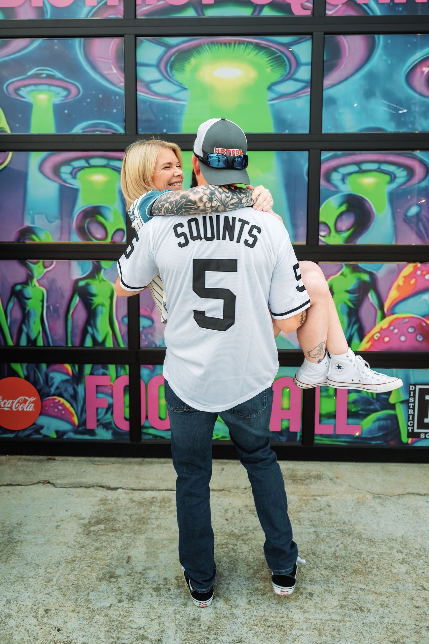 Couple embracing in front of District Square alien mural, man wearing Squints number 5 baseball jersey.