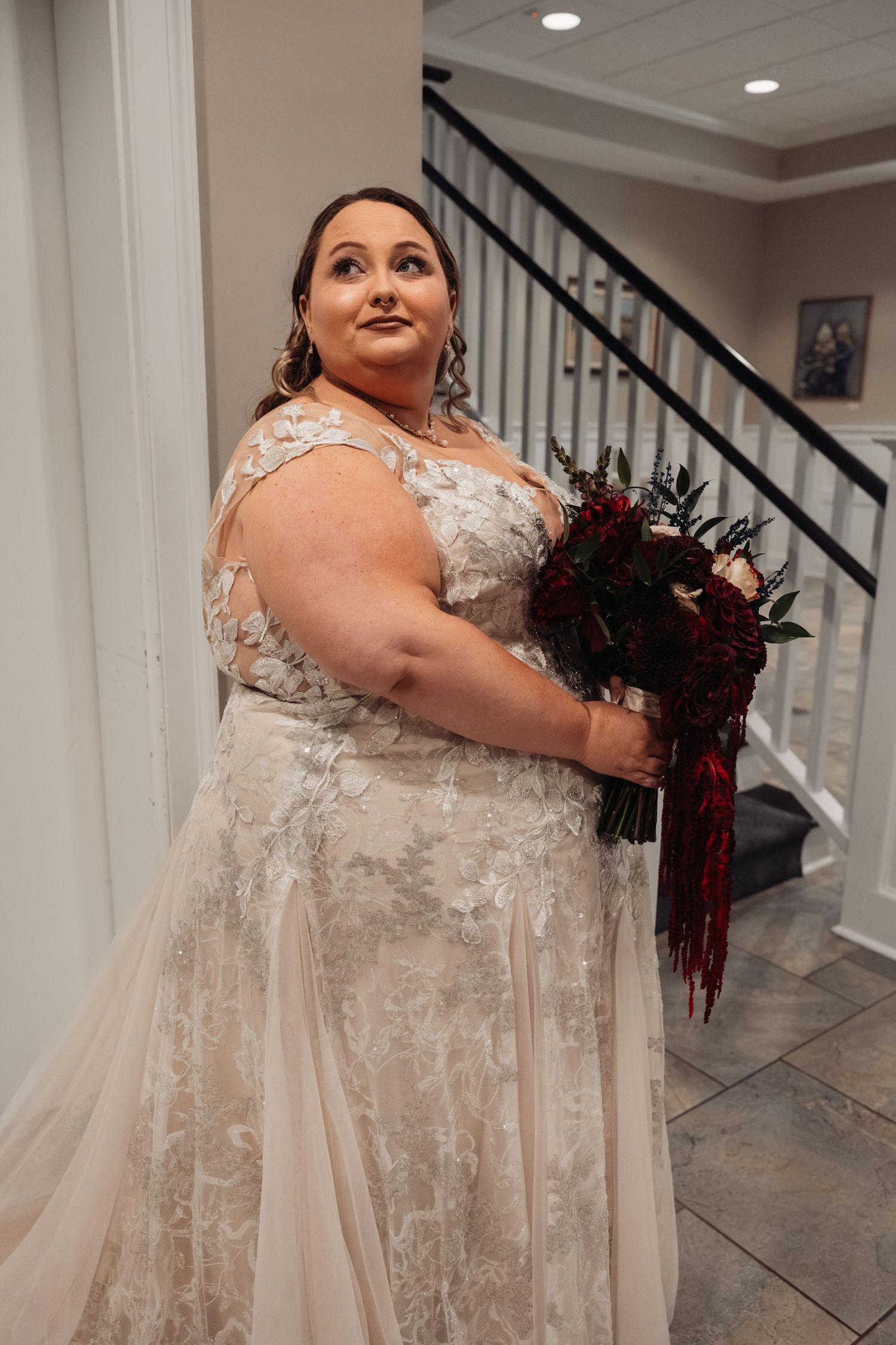 Bride in lace gown holds deep red bouquet on staircase before wedding ceremony.