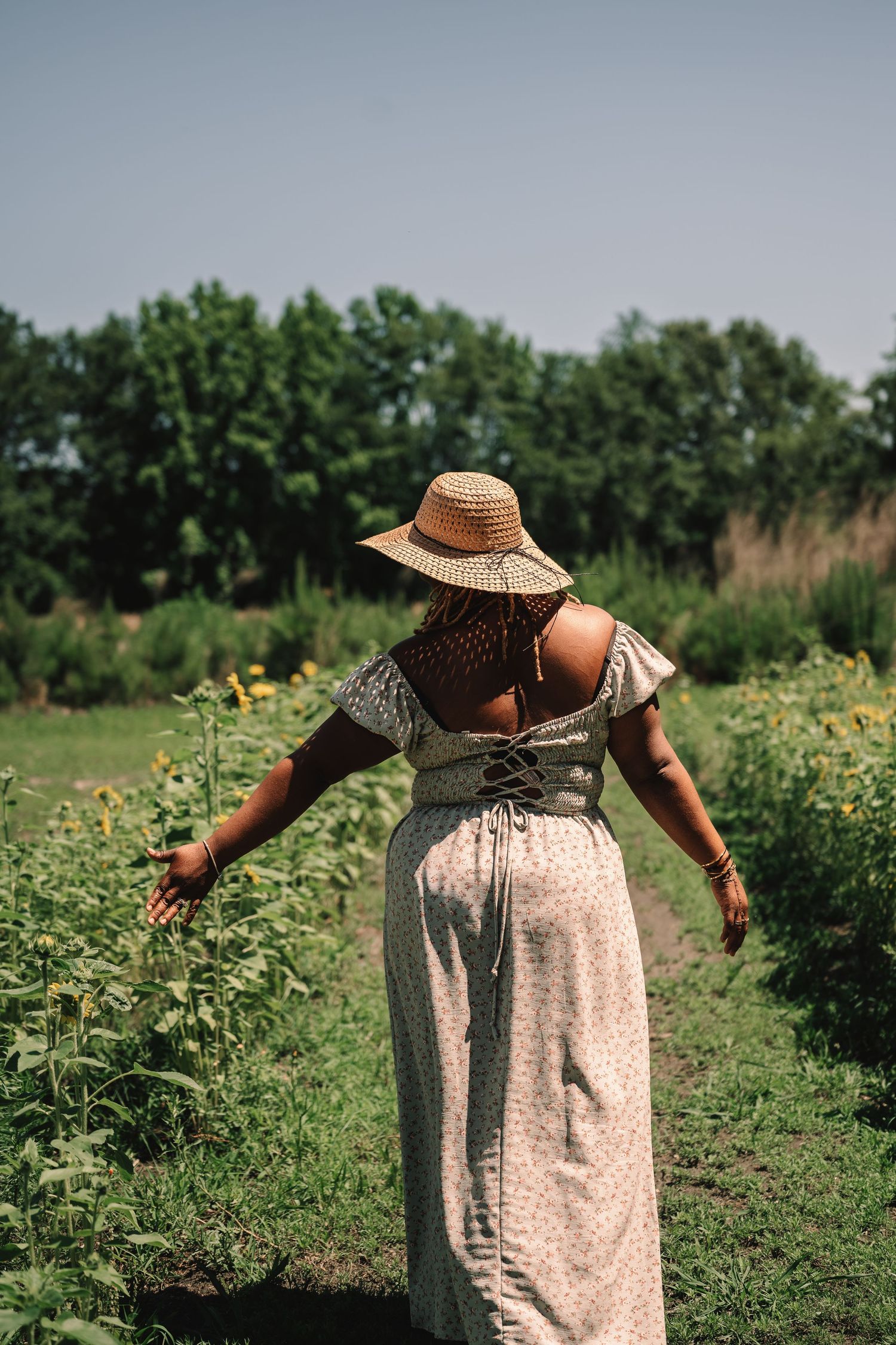 Woman in a floral dress and straw hat walking through a lush sunflower field on a sunny day.