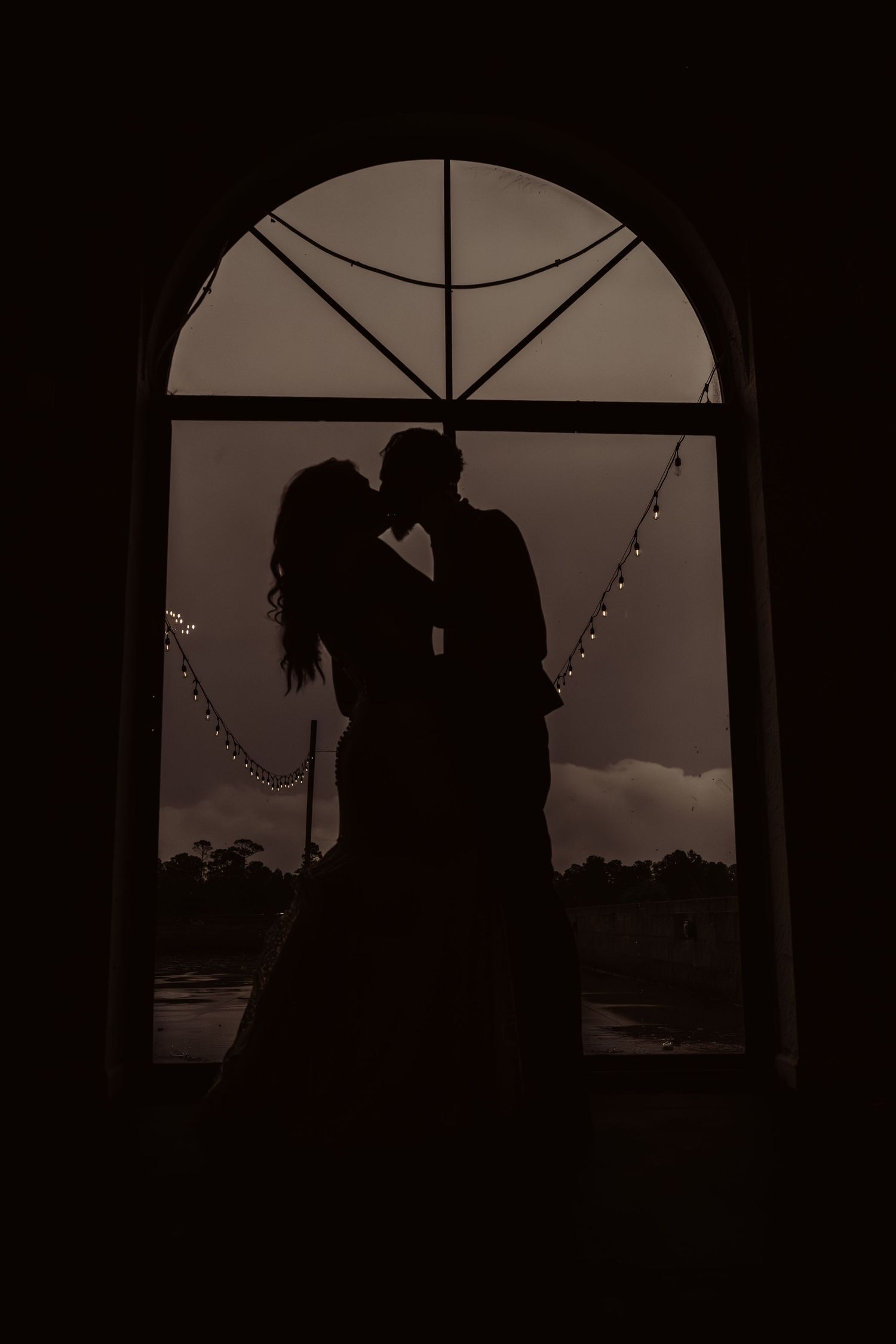 Silhouette of bride and groom kissing in front of an arched window with string lights.