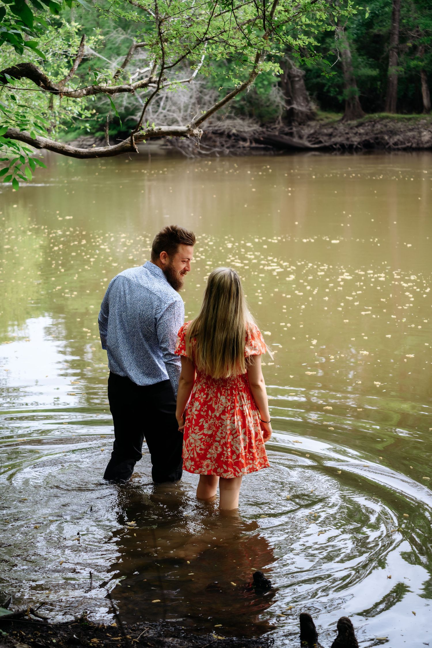 Couple standing together in a shallow river surrounded by lush green trees during an outdoor engagement photo session.