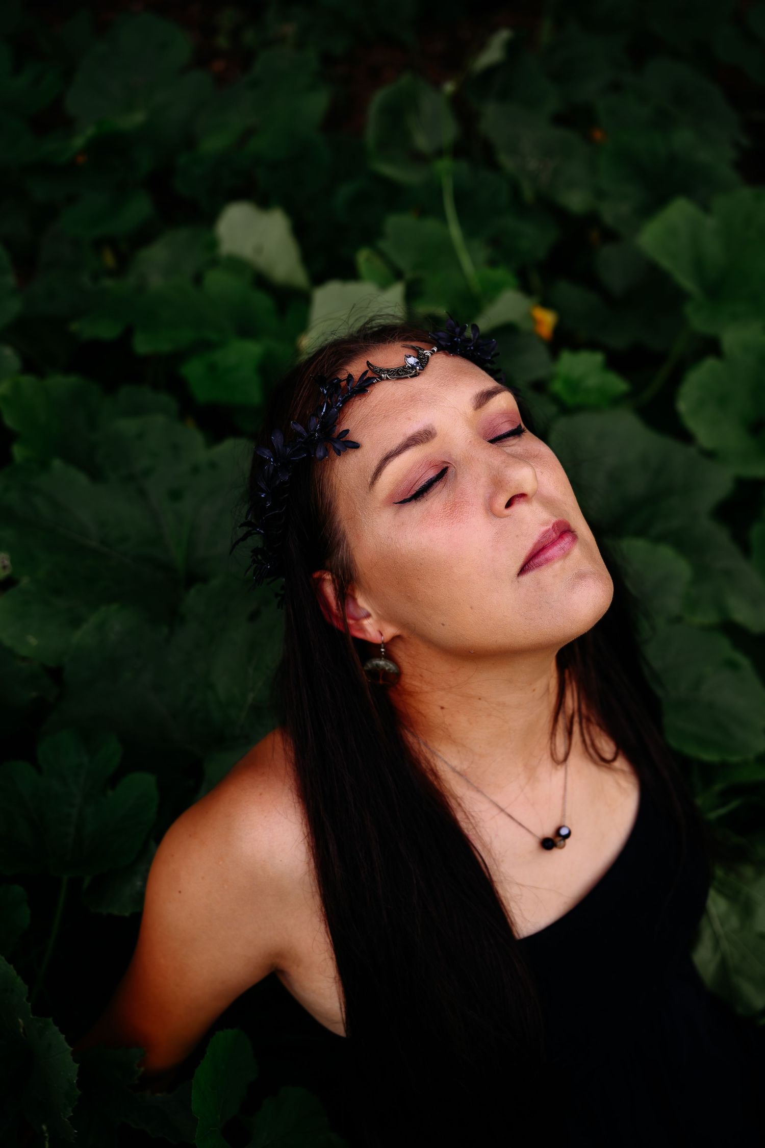 Close-up of woman with black crown headpiece resting among dark green leafy plants.