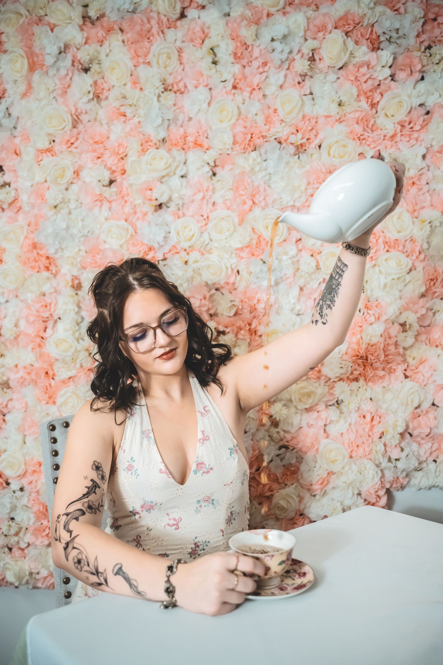 A person in a white dress poses playfully on a bed with pink floral wallpaper background.