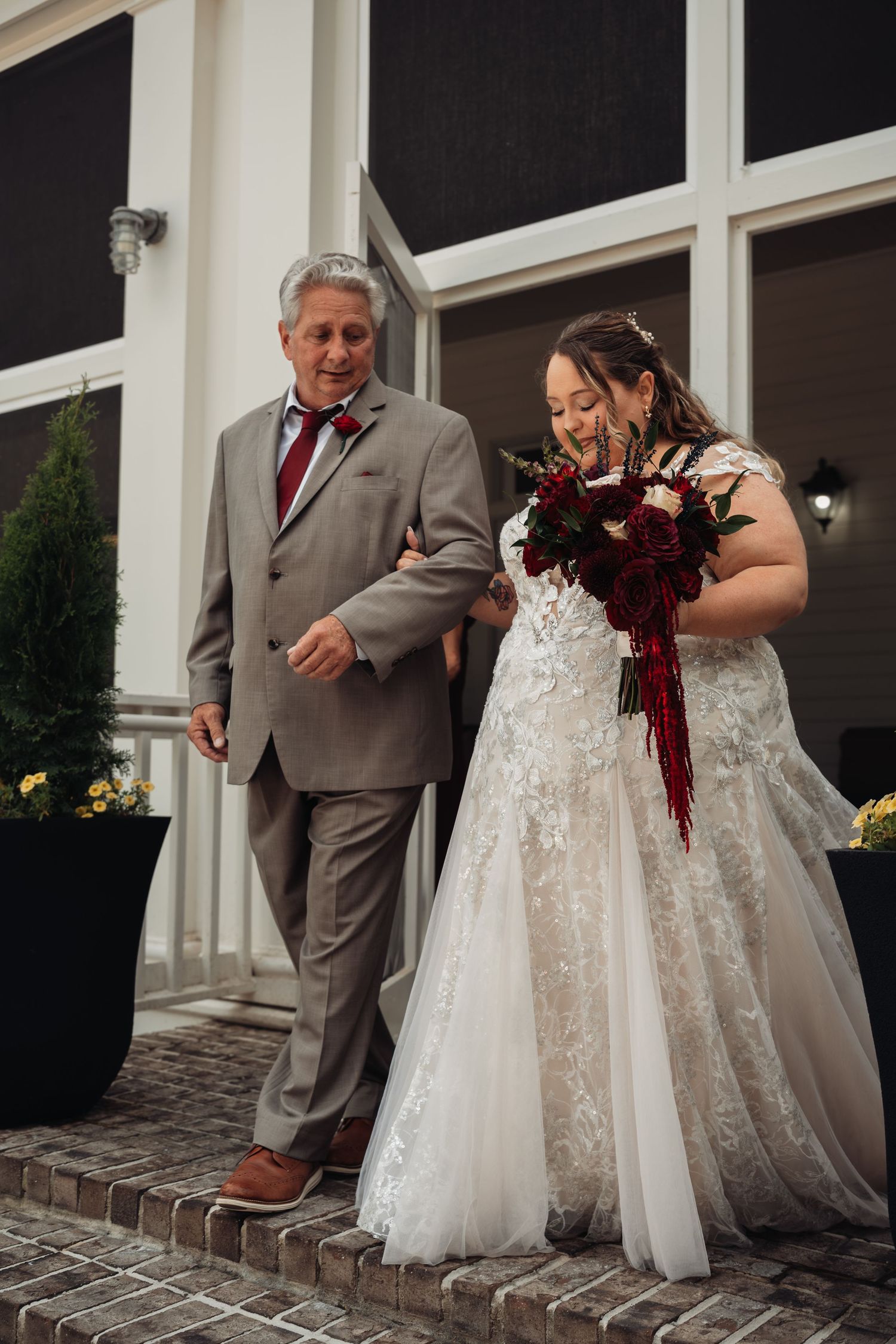 Father escorts bride in lace gown down steps holding deep red floral bouquet.