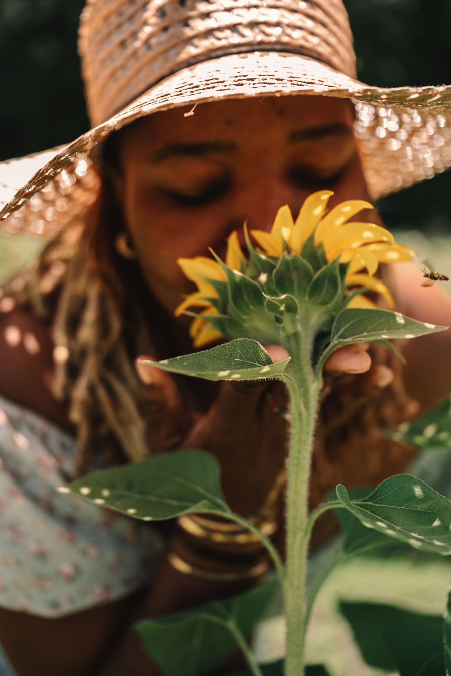 Close-up of a woman in a straw hat leaning in to smell a vibrant yellow sunflower in a garden.