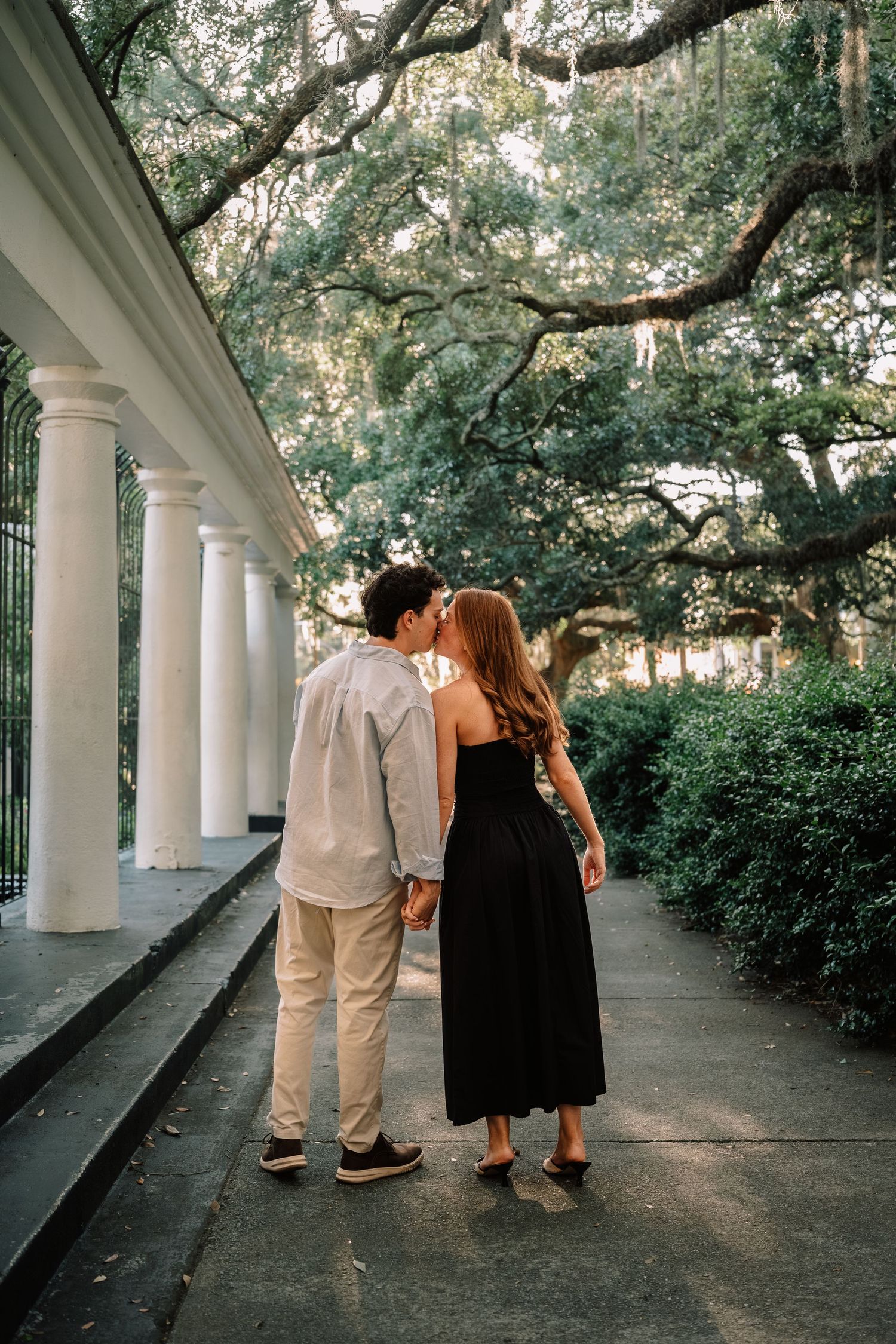 Newly engaged couple sharing a kiss while holding hands near white columns and lush trees.