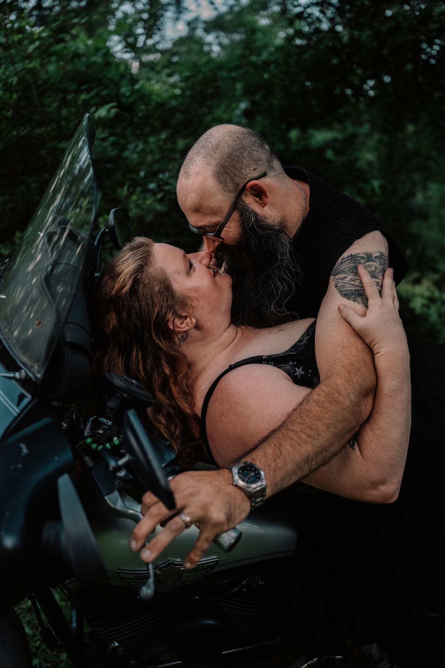 Bearded tattooed man and woman sharing an intimate moment on a motorcycle outdoors.