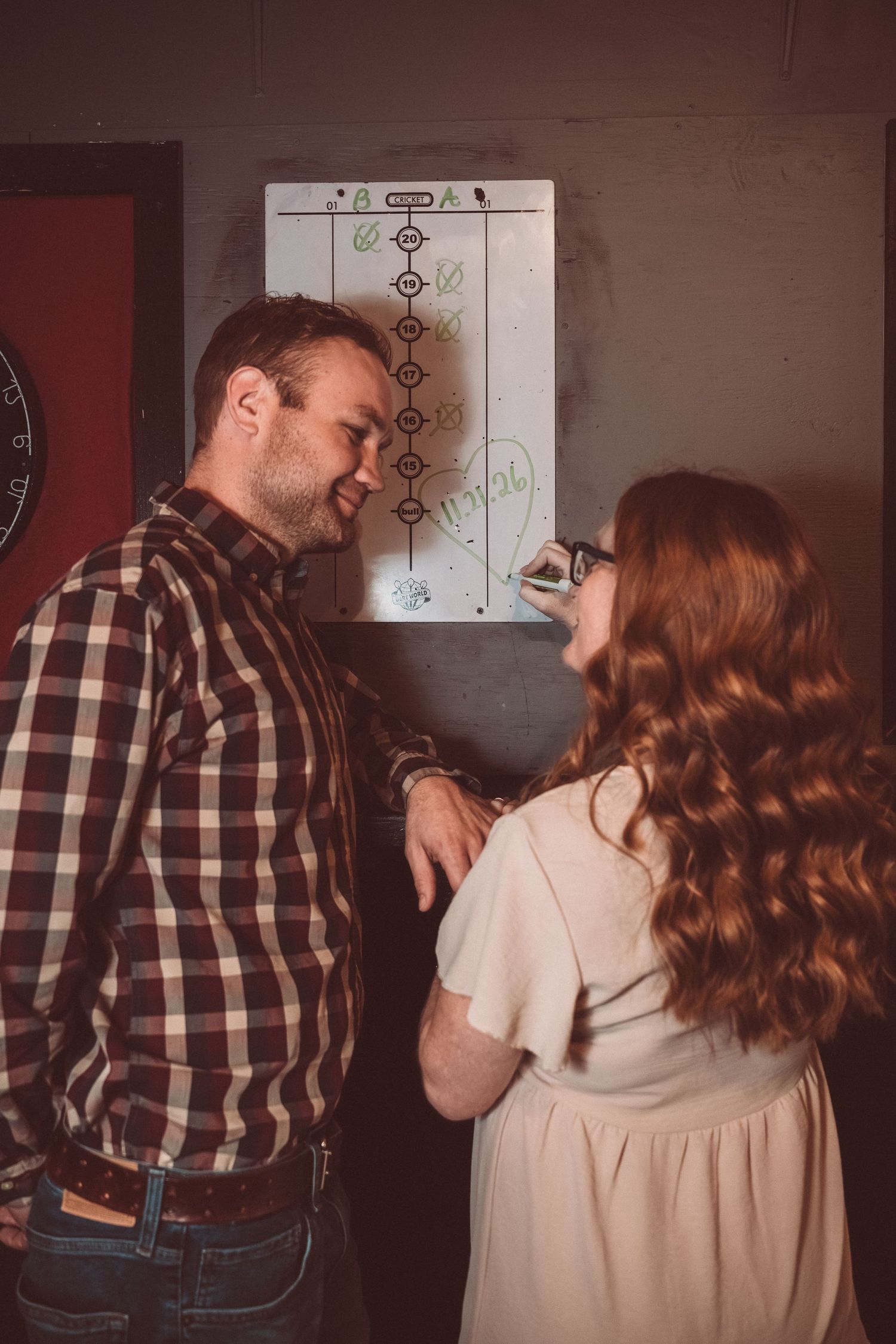 A couple shares a close moment next to a strategy board hanging on the wall in warm lighting.