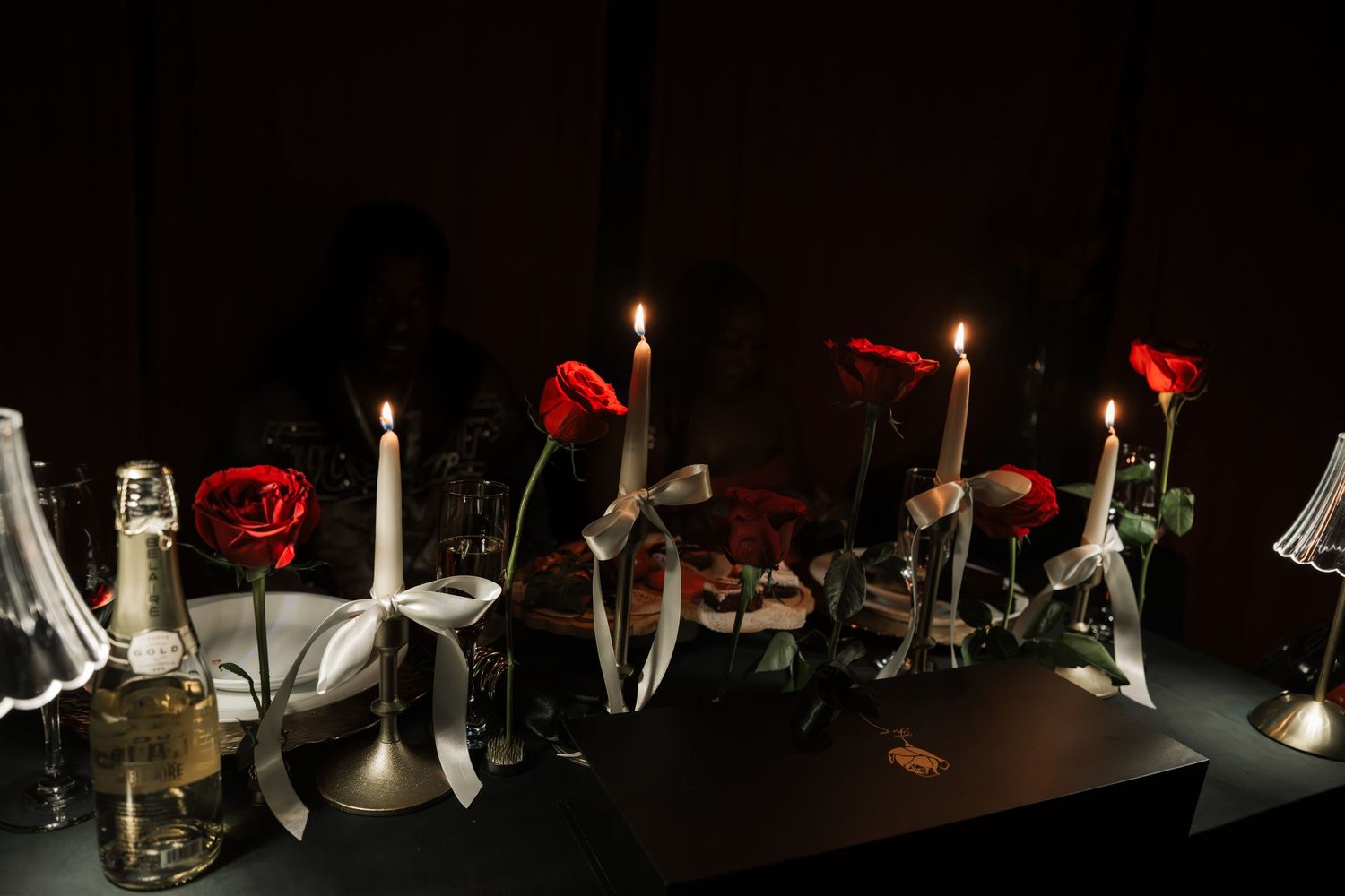 Elegant candlelit table decorated with red roses and white ribbon centerpieces in dark romantic setting.