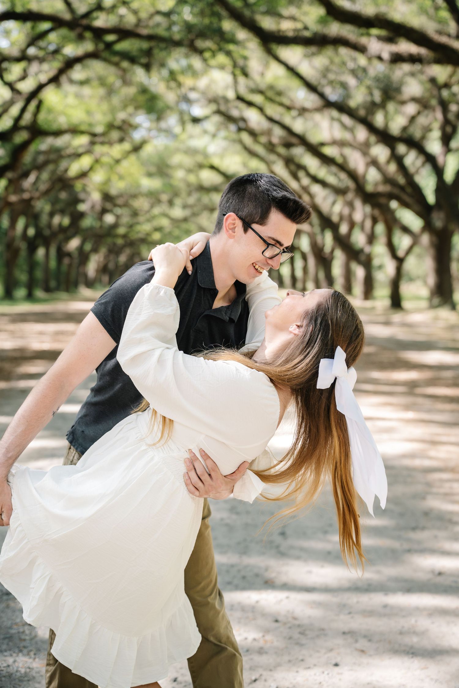 Man dipping woman backward in a romantic pose along a tree-lined path in a park.