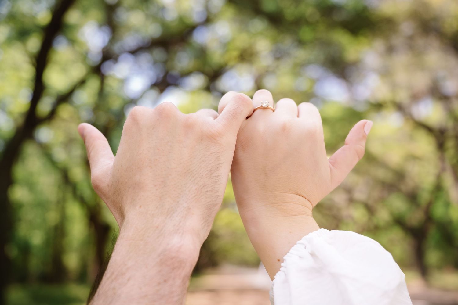 Two people making a pinky promise with an engagement ring visible, set against a lush green park background.