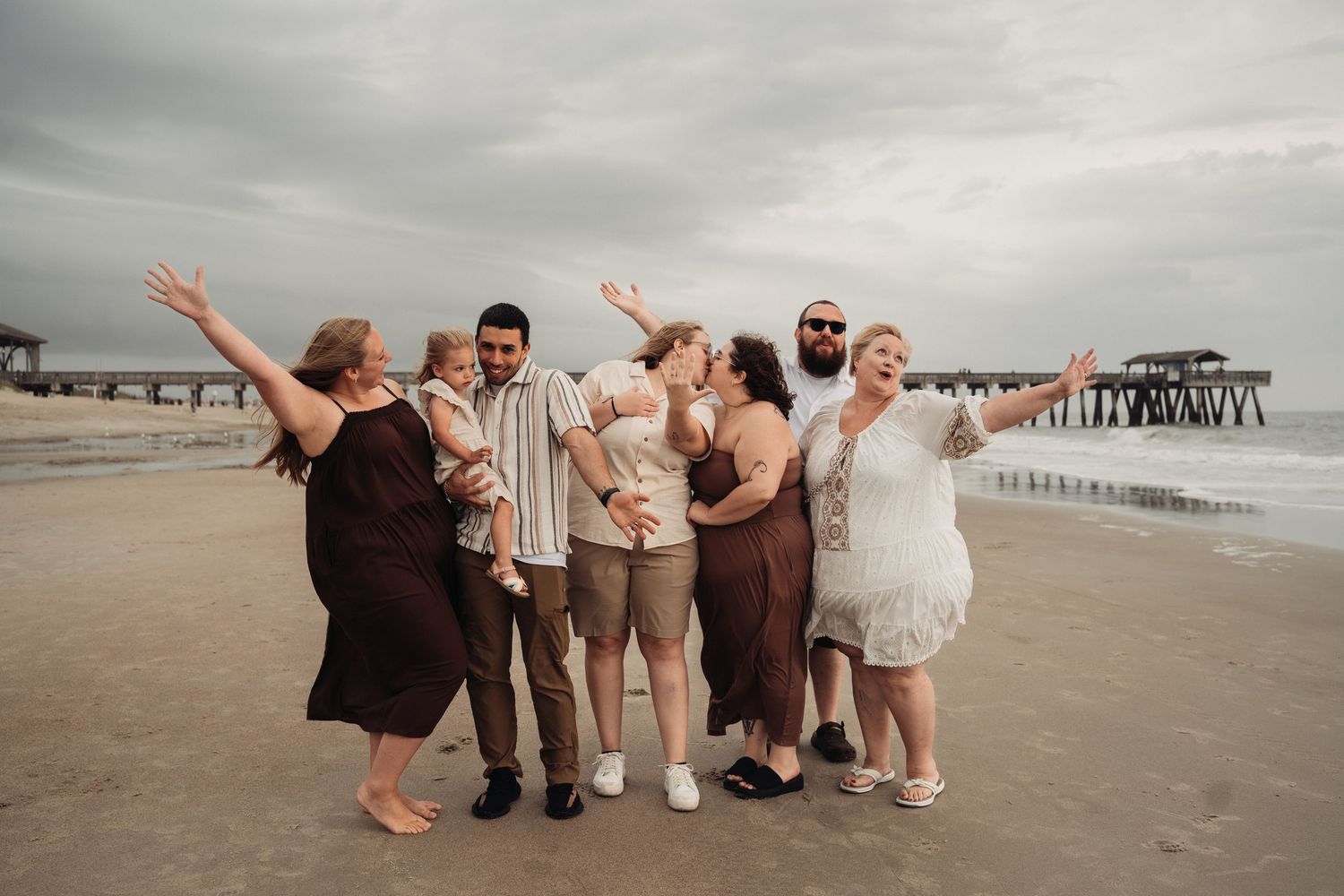 A joyful group of six people posing and celebrating on a beach near a wooden pier under cloudy skies.