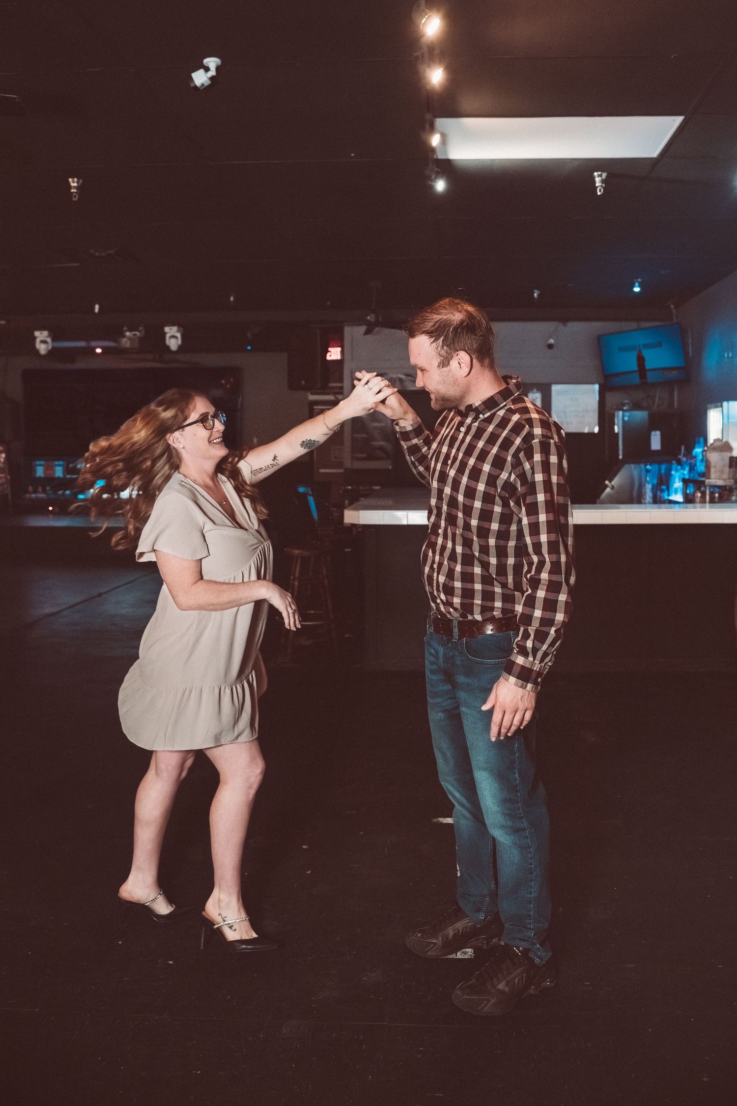 Two people dancing together in a dimly lit venue, with one wearing a plaid shirt and the other in a light-colored dress.