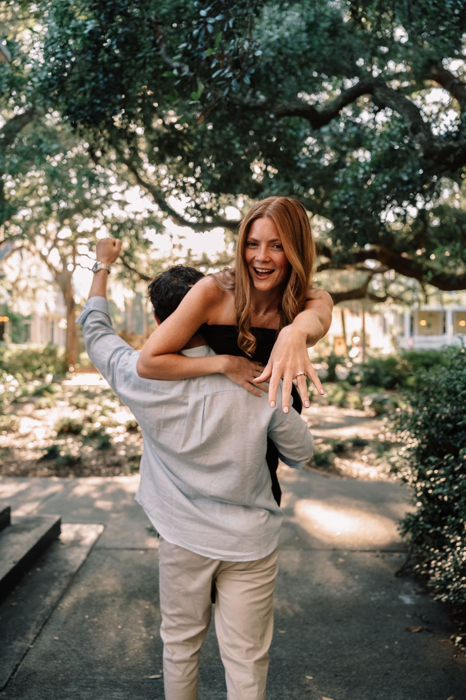 Joyful woman showing engagement ring while being lifted by her fiance in a lush park.