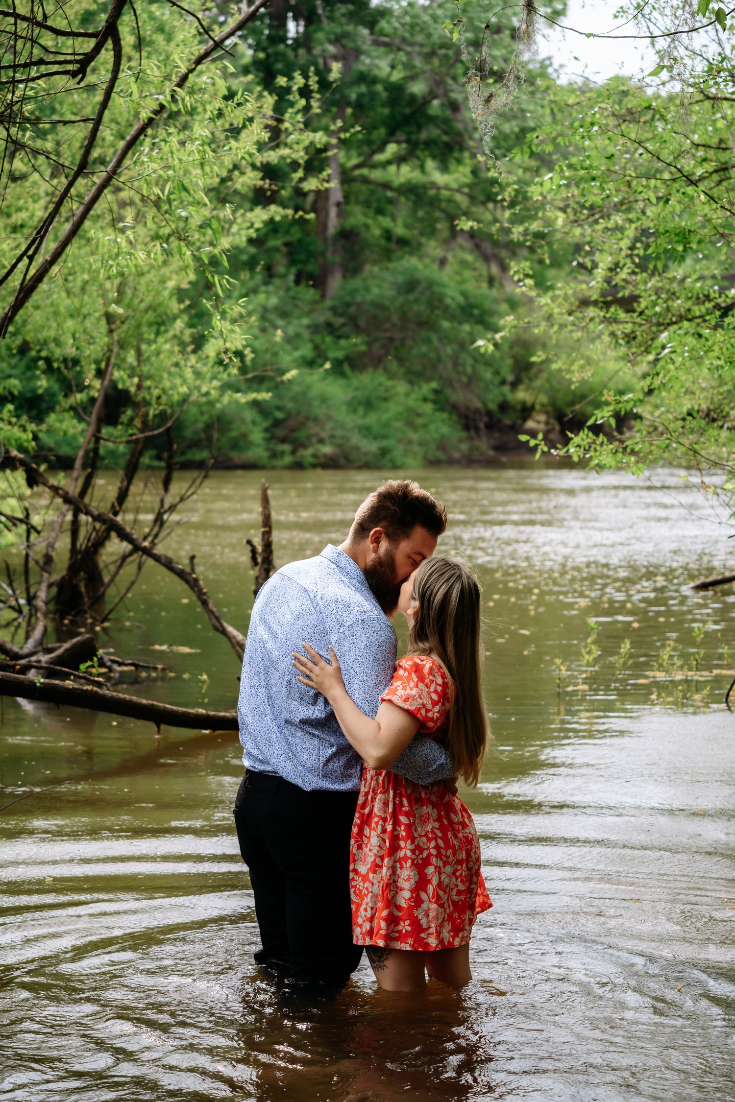 Couple sharing a romantic kiss while standing in a shallow creek surrounded by lush green trees.