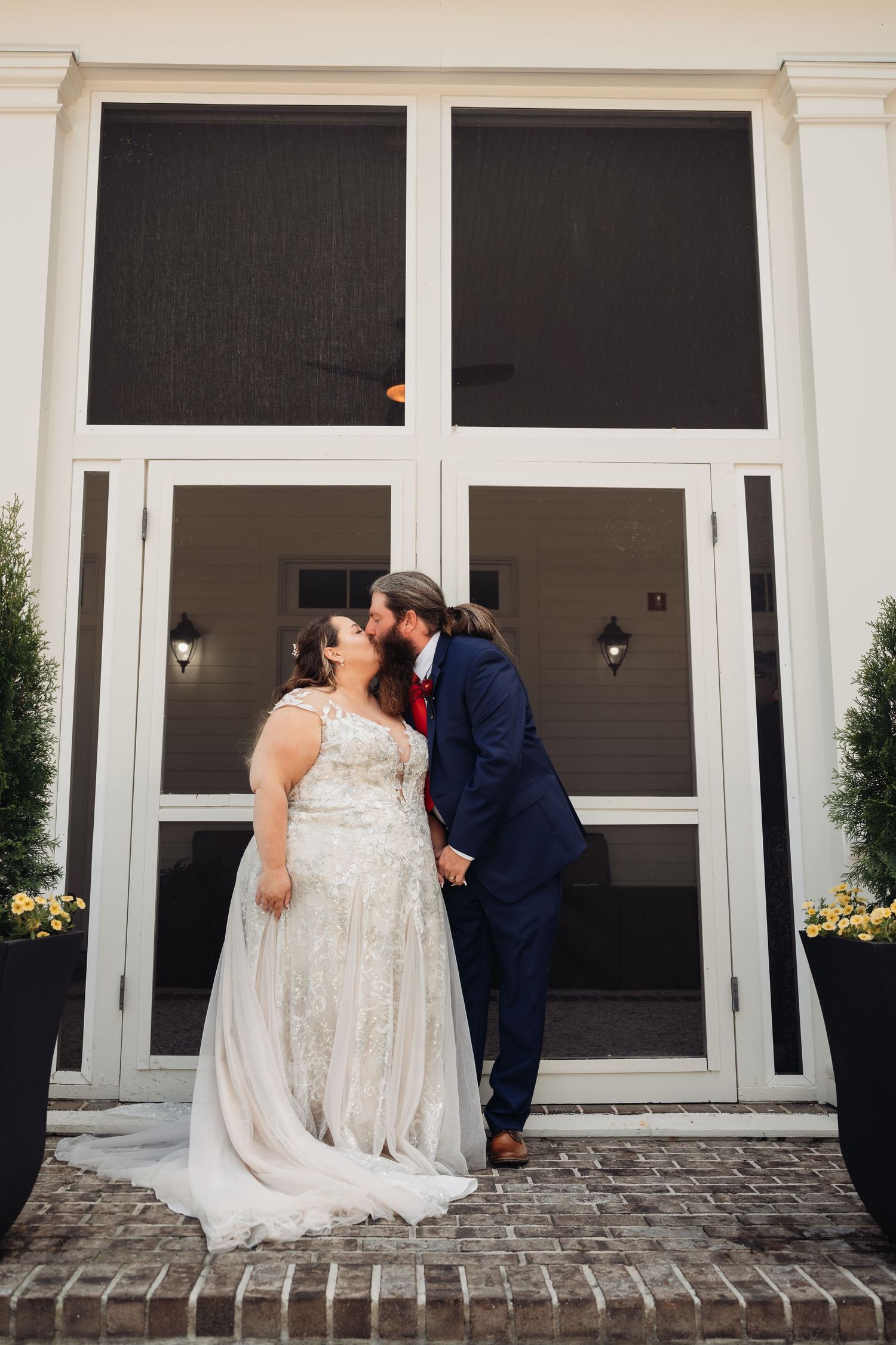 Groom in navy suit kisses bride in lace gown outside white wedding venue.