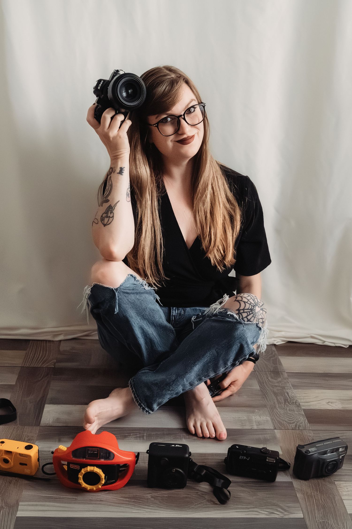 Female photographer sitting cross-legged on floor holding camera surrounded by vintage cameras.