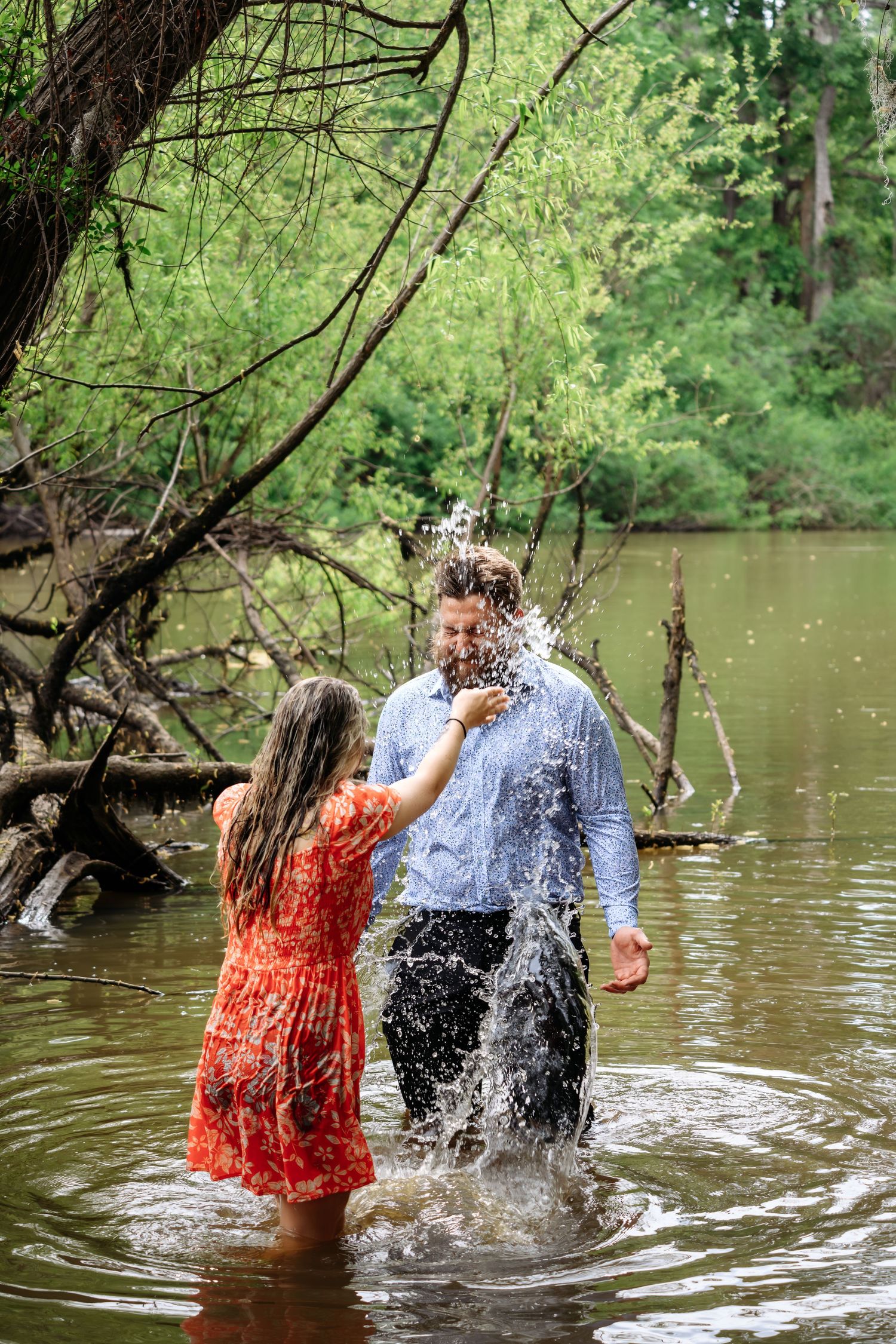Couple splashing water in a river, woman in red dress and man in blue shirt, surrounded by lush green trees.