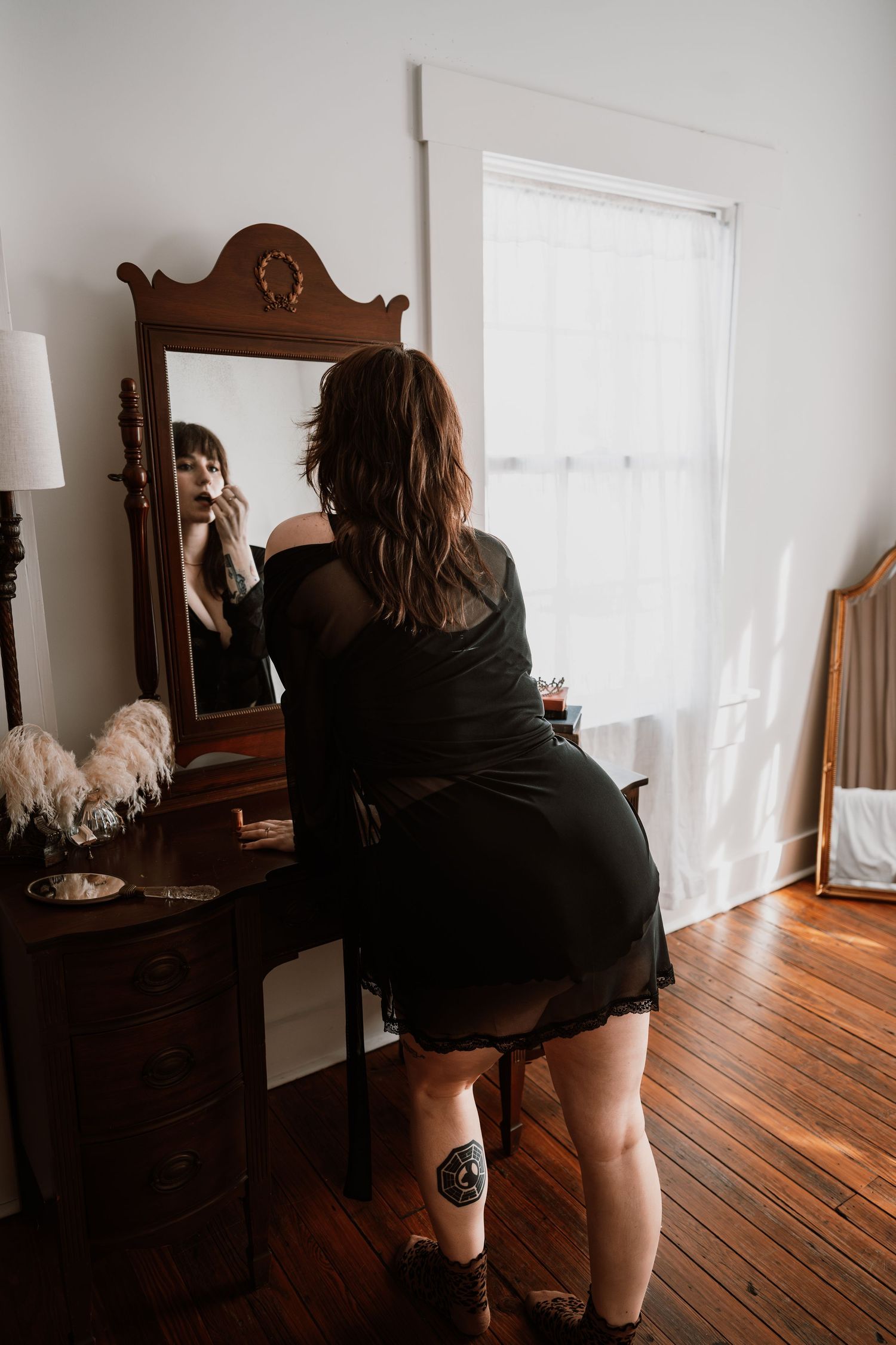 Woman in black dress applies makeup at antique vanity mirror in vintage bedroom.