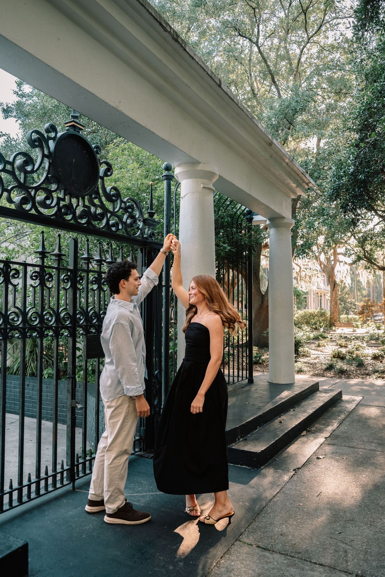 Couple dancing near ornate iron gate and white columns surrounded by green trees outdoors.