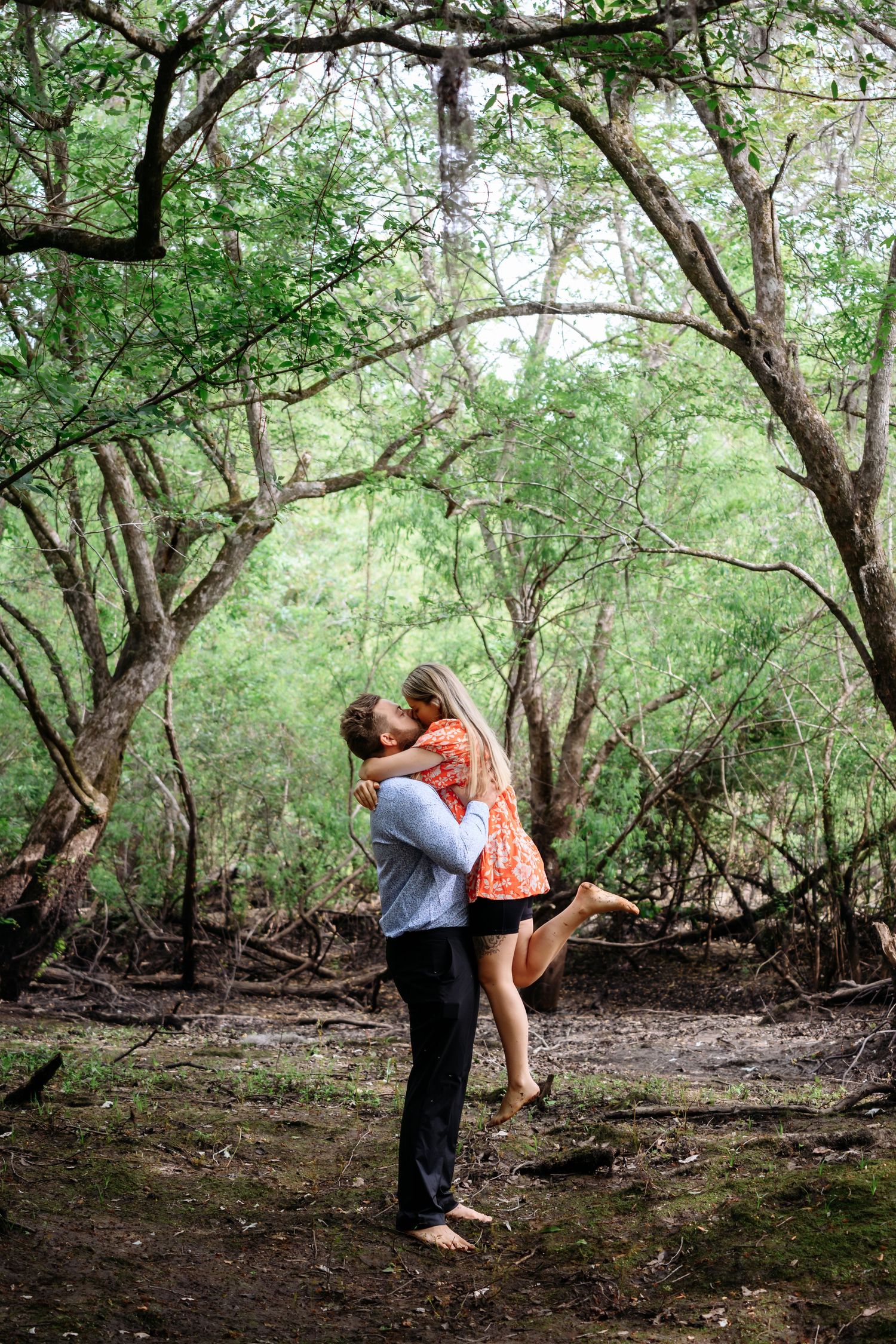 Couple sharing a romantic kiss in a lush green forest, man lifting woman in his arms.